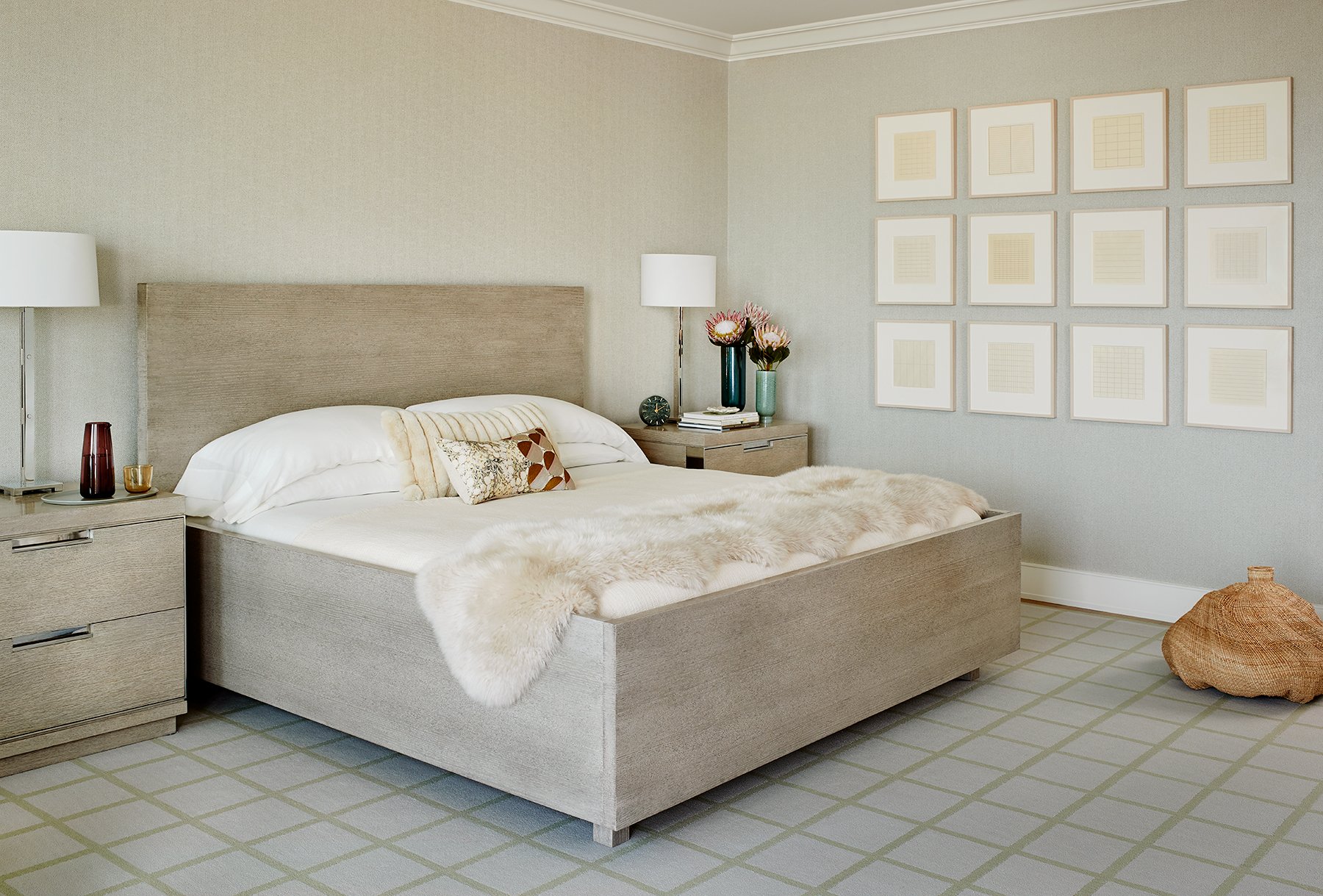 A bedroom with a beige upholstered bed, white bedding, decorative pillows, two nightstands with lamps, flowers, and books, a wall with framed artwork, a beige and green checkered carpet, and a wicker basket on the floor on Filbert Pacific Height