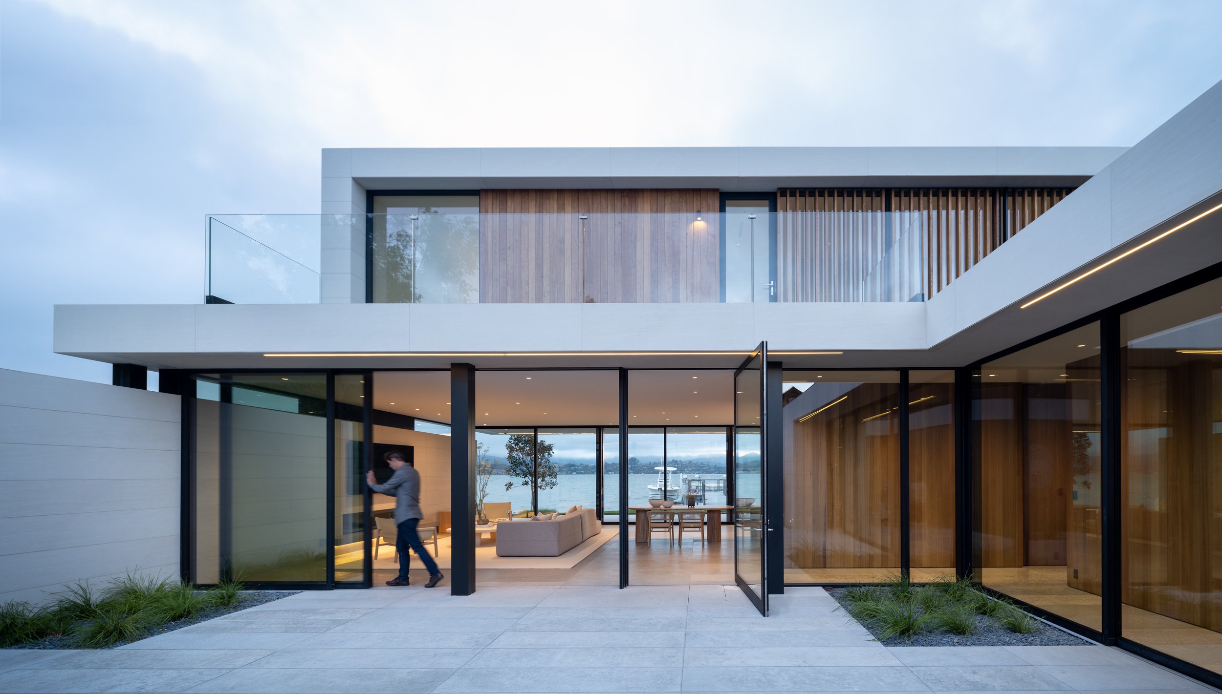 Modern two-story house with large glass windows and sliding doors, overlooking a body of water with boats, featuring a patio with minimalist outdoor furniture and a man walking inside.  Interior Design by Ballon Studio and Architecture by Bulter Arms