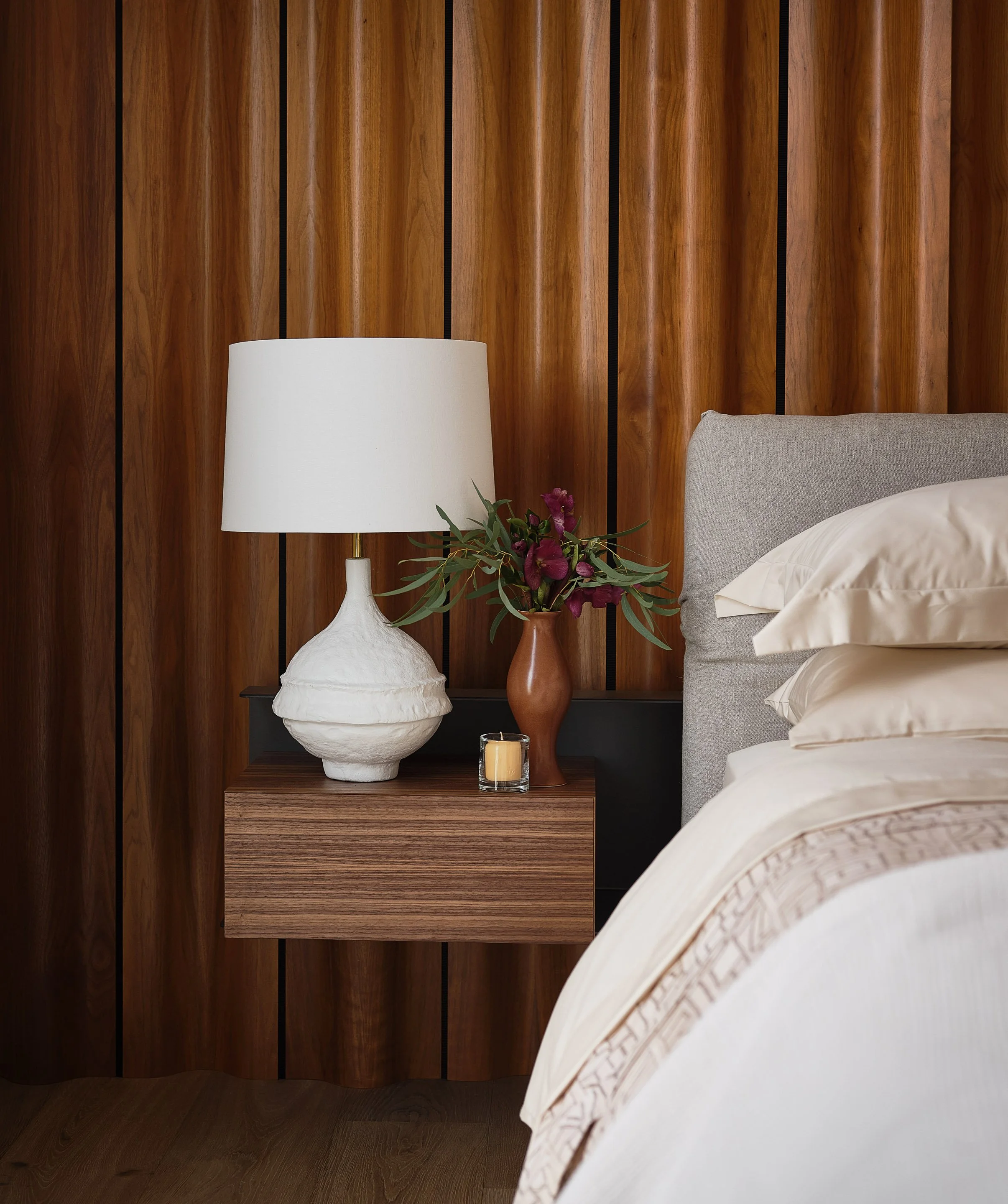 Broadway Penthouse Pacific Heights bedside vignette with a sculptural white lamp, floral arrangement, and candle beside layered neutral bedding, set against a warm wood paneled wall for calm modern luxury in San Francisco.