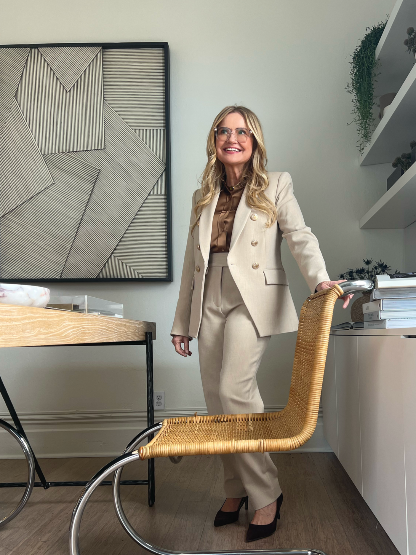 Sabra Ballon in the ballonSTUDIO office in 2025, smiling in a beige suit beside a rattan chair, framed by modern art and styled shelves with plants, reflecting a refined San Francisco interior design studio.
