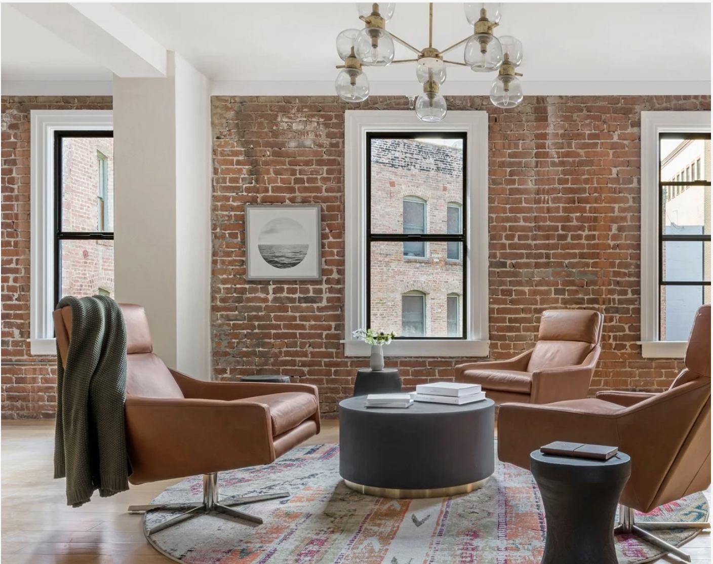 Geometer tech office with an exposed brick wall and three large windows, styled with brown leather lounge chairs, a round black coffee table with books, and a modern glass globe chandelier over a patterned rug.