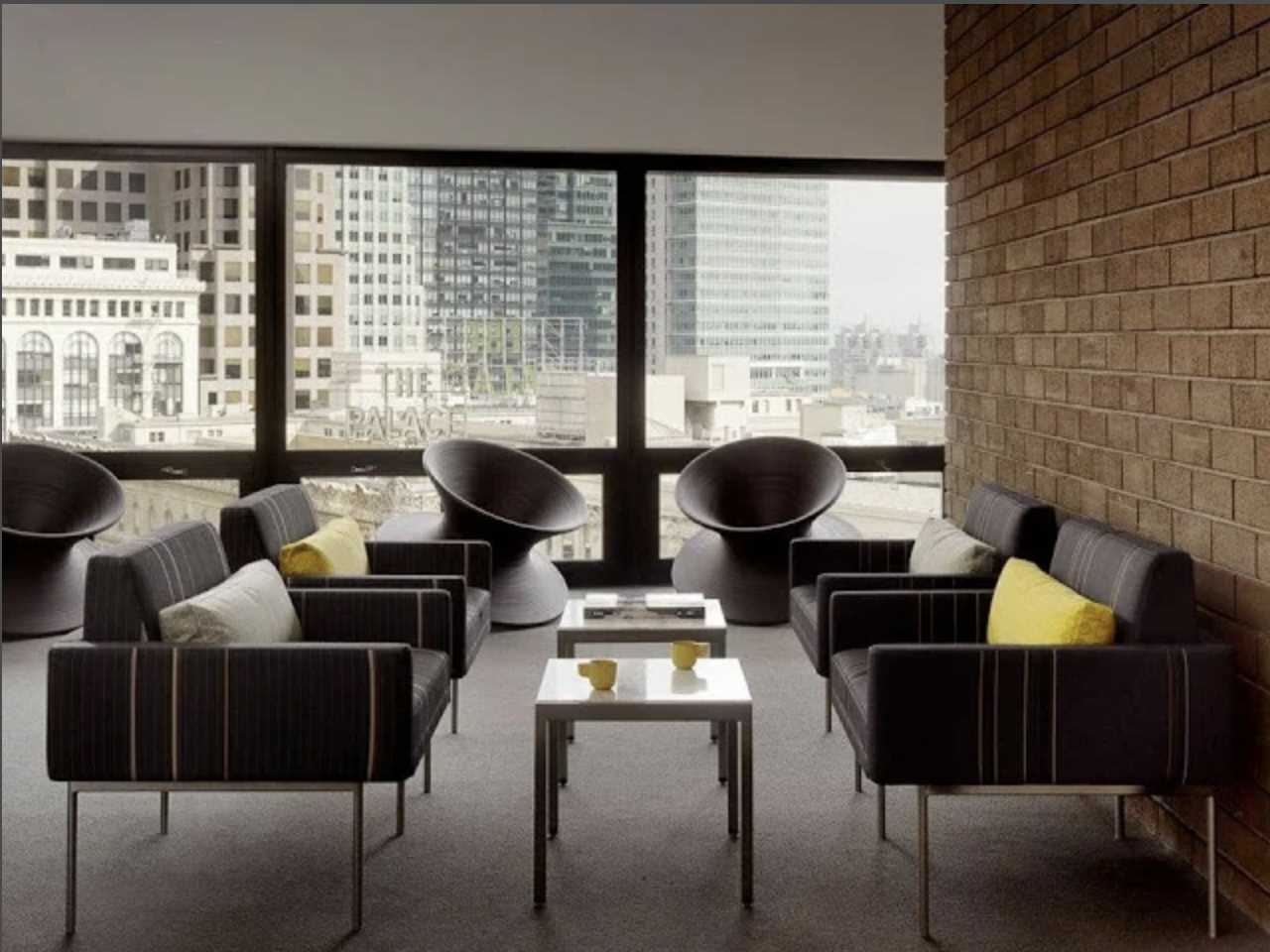 Modern hotel lounge with black armchairs and yellow accent pillows, styled with white side tables and yellow cups, framed by large windows overlooking downtown skyscrapers for a bright, urban hospitality setting.