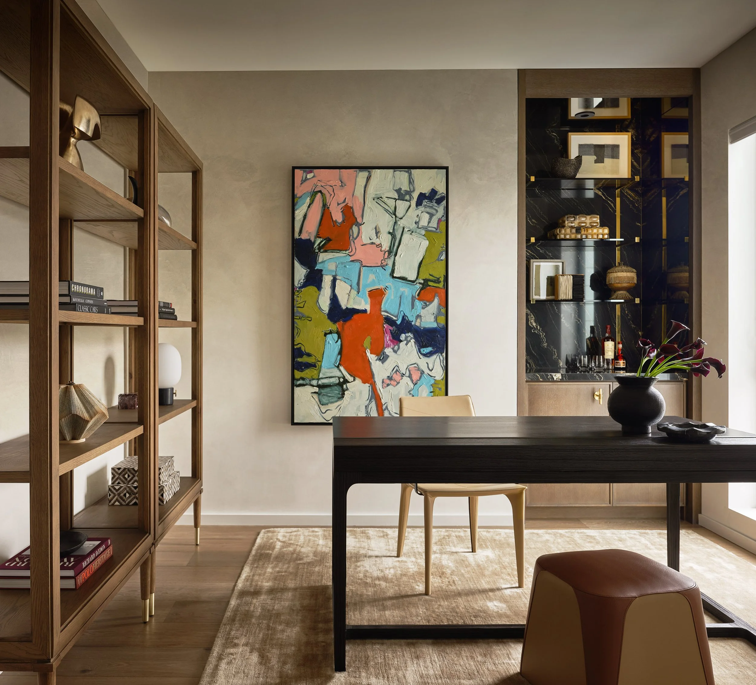 Art consultant with white glove installation service for Broadway Penthouse home office with a wooden bookshelf, abstract art painting, black marble wall with gold accents, dining table with a vase of dark purple flowers, and a beige rug.