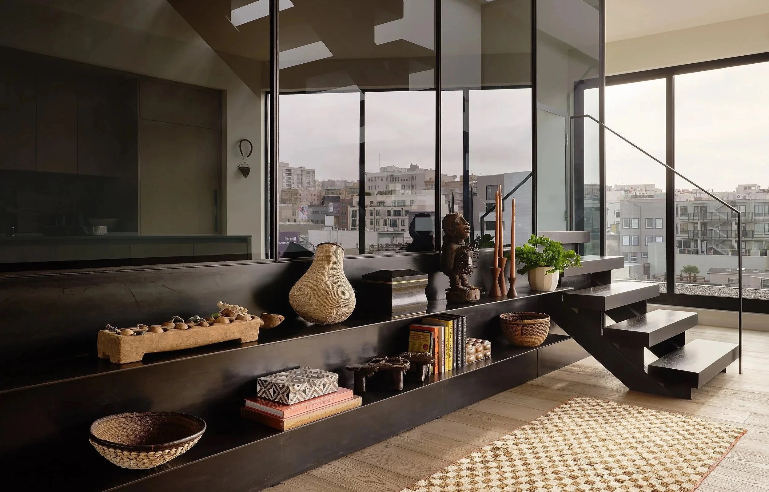 Interior of a modern home with a black staircase and a black built-in shelf adorned with various decorative objects, books, and plants. Large windows reveal a cityscape outside.