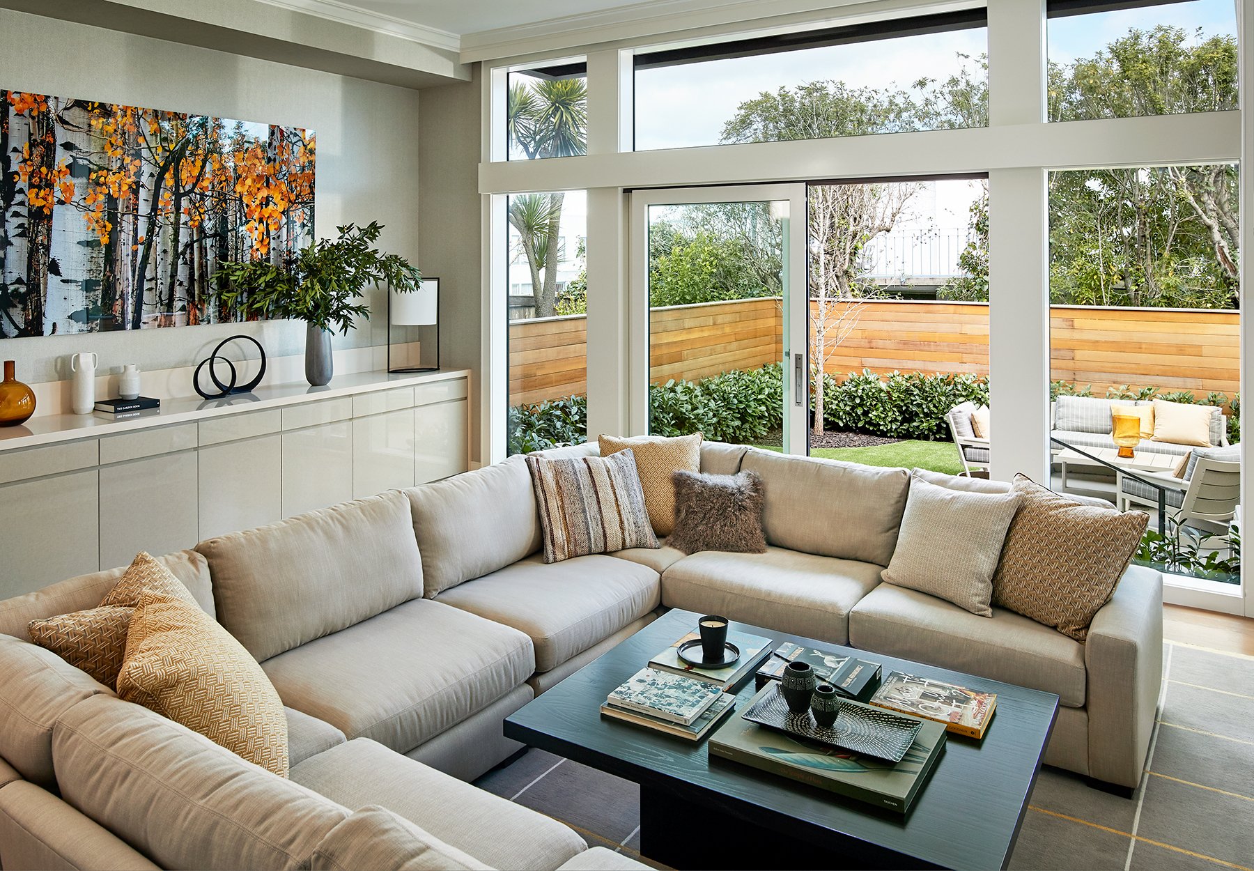Modern living room with large windows, beige sectional sofa with throw pillows, coffee table with magazines and decorative items, and outdoor patio with seating and greenery visible through the windows on Filbert Street Pacific Heights in San Fran.