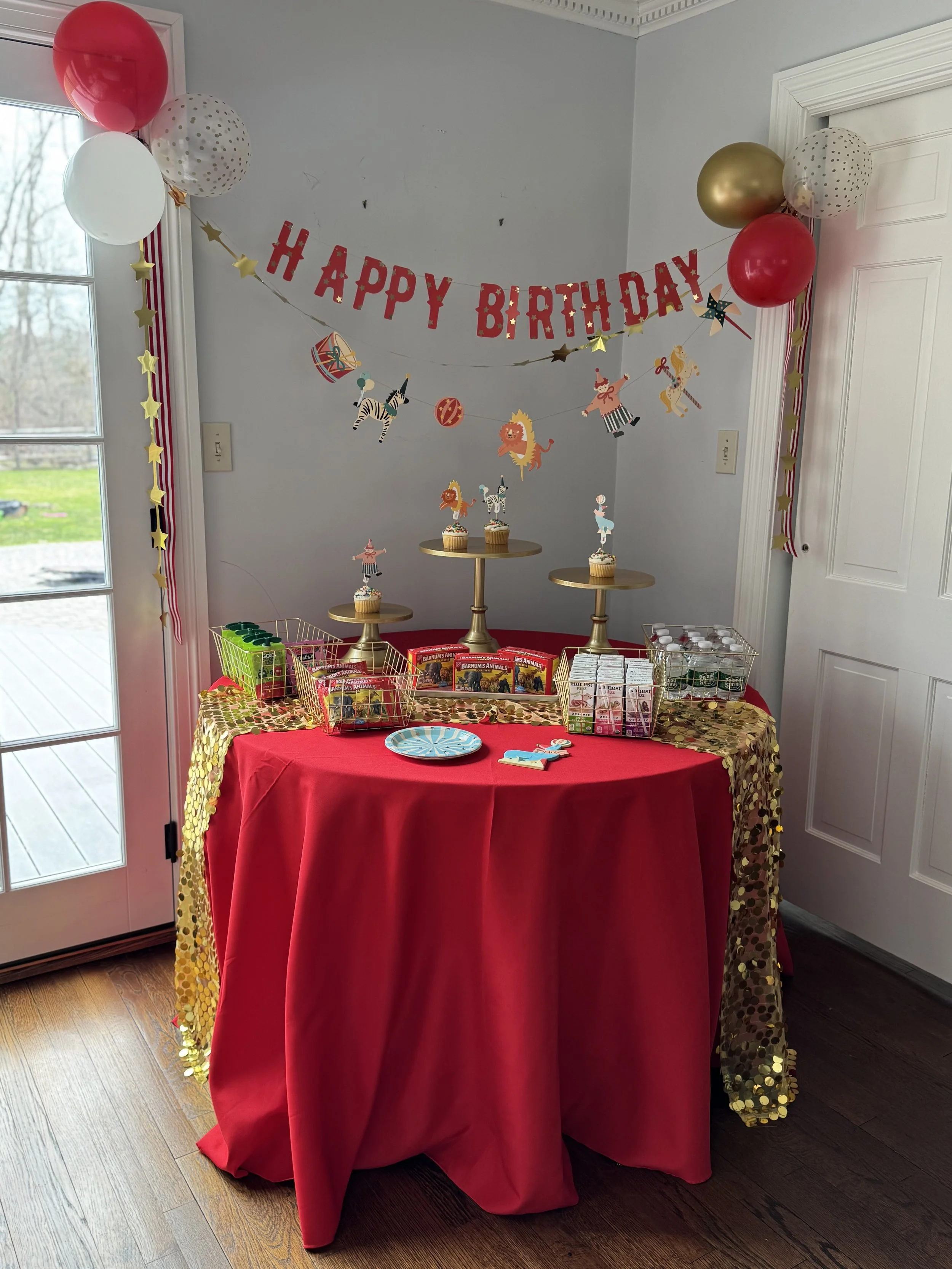 A birthday party setup with a red tablecloth, gold sequin table runner, birthday cake cupcakes with animal toppers, and a 'Happy Birthday' banner with circus animal decorations in the background.