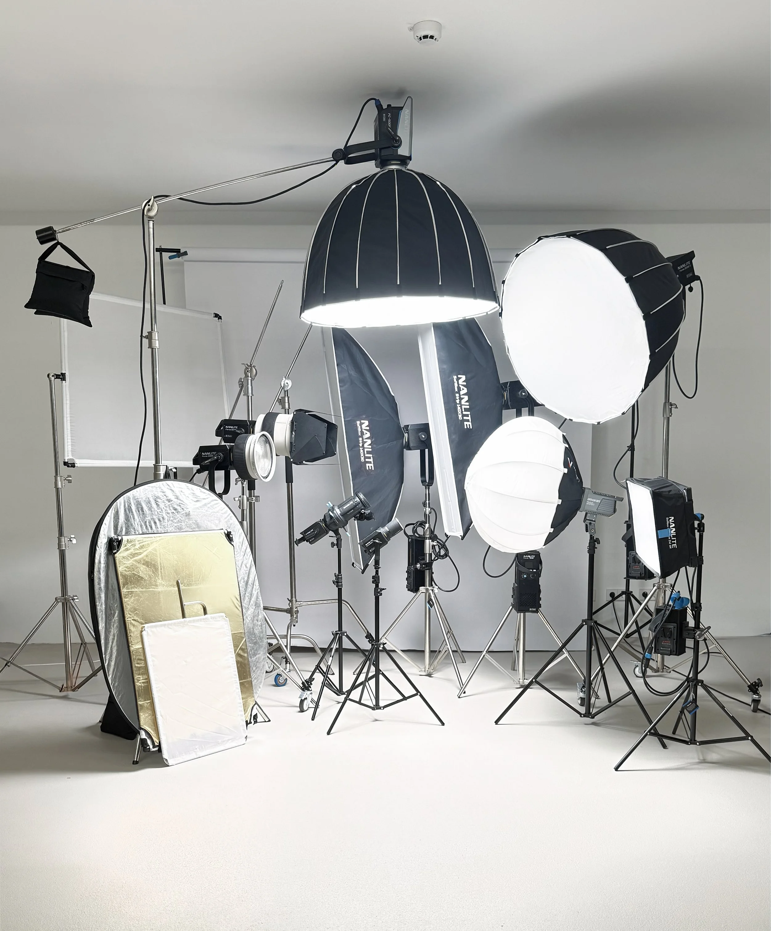 Photography studio setup with various lighting equipment, softboxes, reflectors, and backdrop stands in a whiteroom.