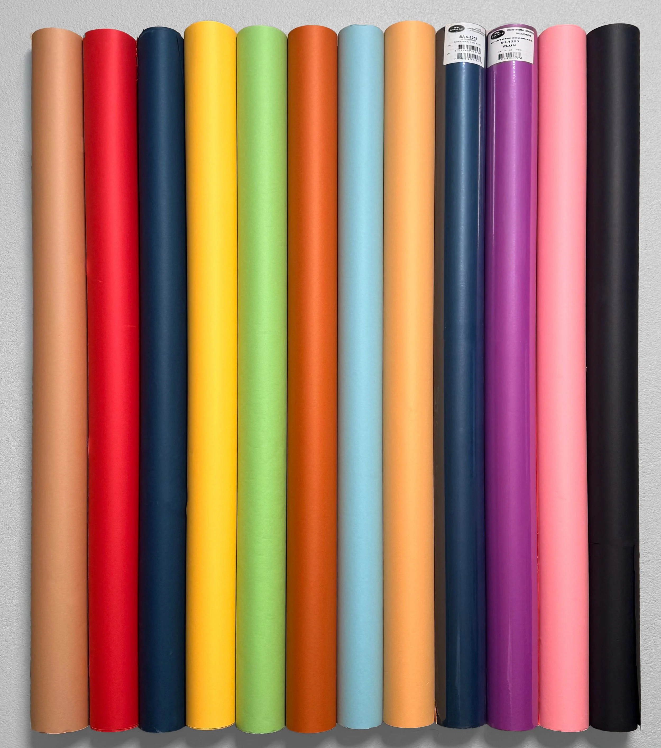 A row of colorful wrapping paper rolls arranged vertically on a light gray surface, featuring colors such as beige, red, navy blue, yellow, green, orange, light blue, peach, purple, pink, and black.