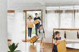 A family of four with two children entering a home carrying luggage, with a woman and man in the background.