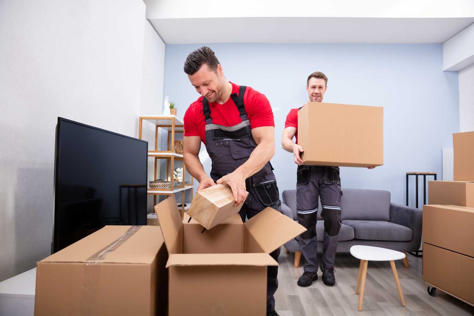 Two men in red shirts and work overalls unpacking boxes and carrying moving boxes in a living room.