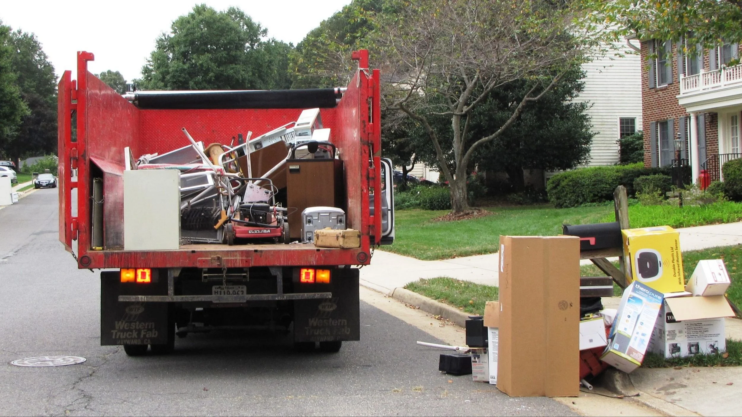 Red trash truck parked on a suburban street with household items and boxes filled with discarded items nearby.