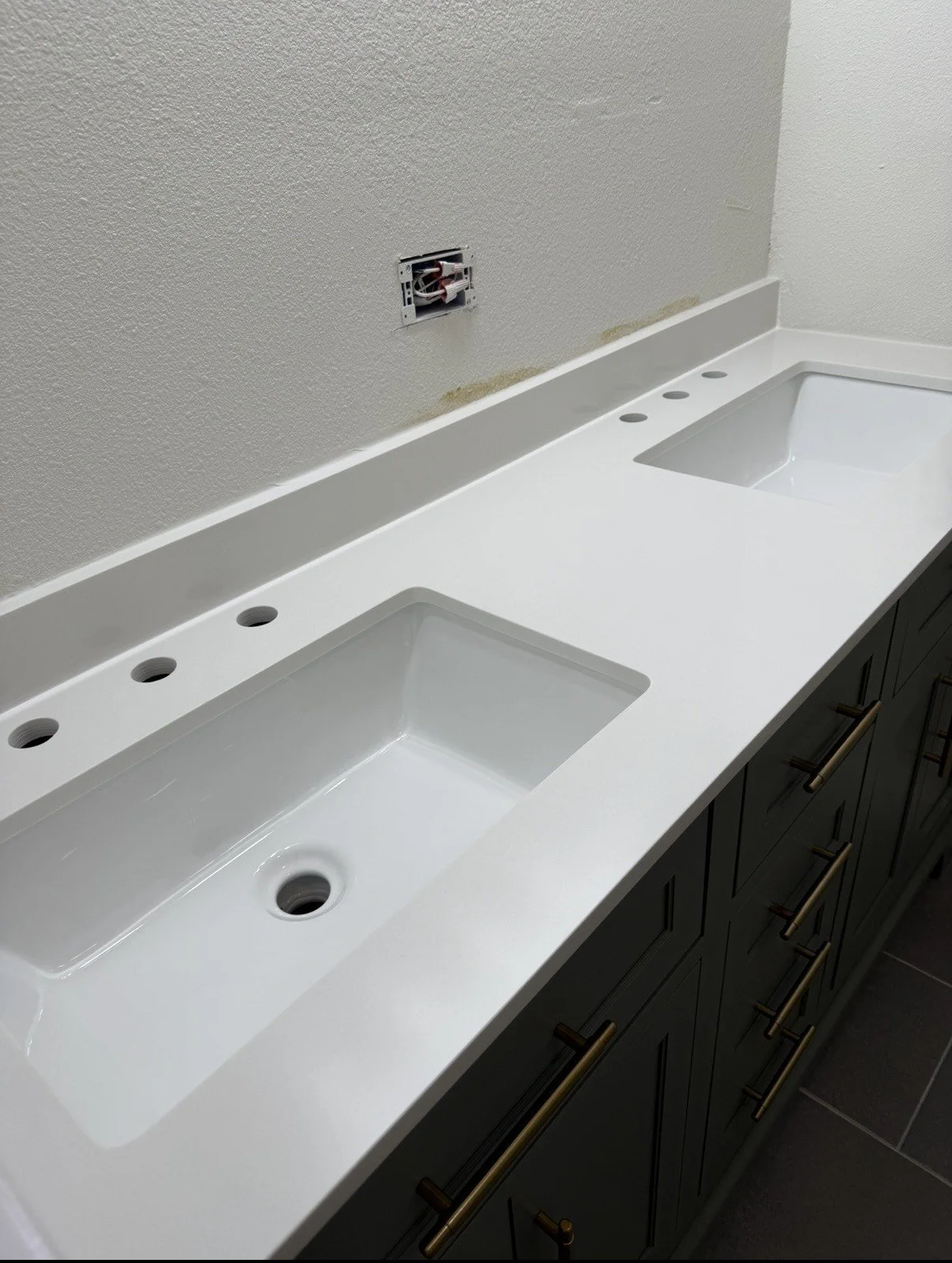 A double bathroom vanity with a white countertop and two rectangular undermount sinks, with six faucet holes, dark cabinetry with gold handles, and an unfinished wall with an electrical outlet.