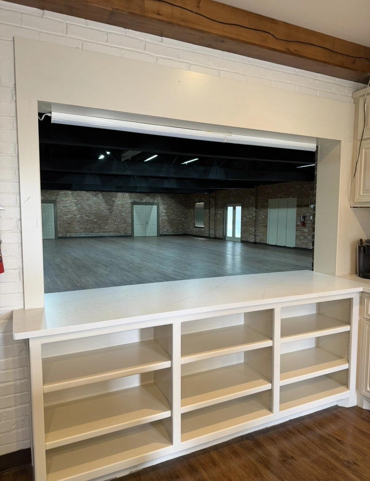 View of an empty room with exposed brick walls, large windows, and sloped ceiling, seen through a kitchen pass-through with built-in shelves.