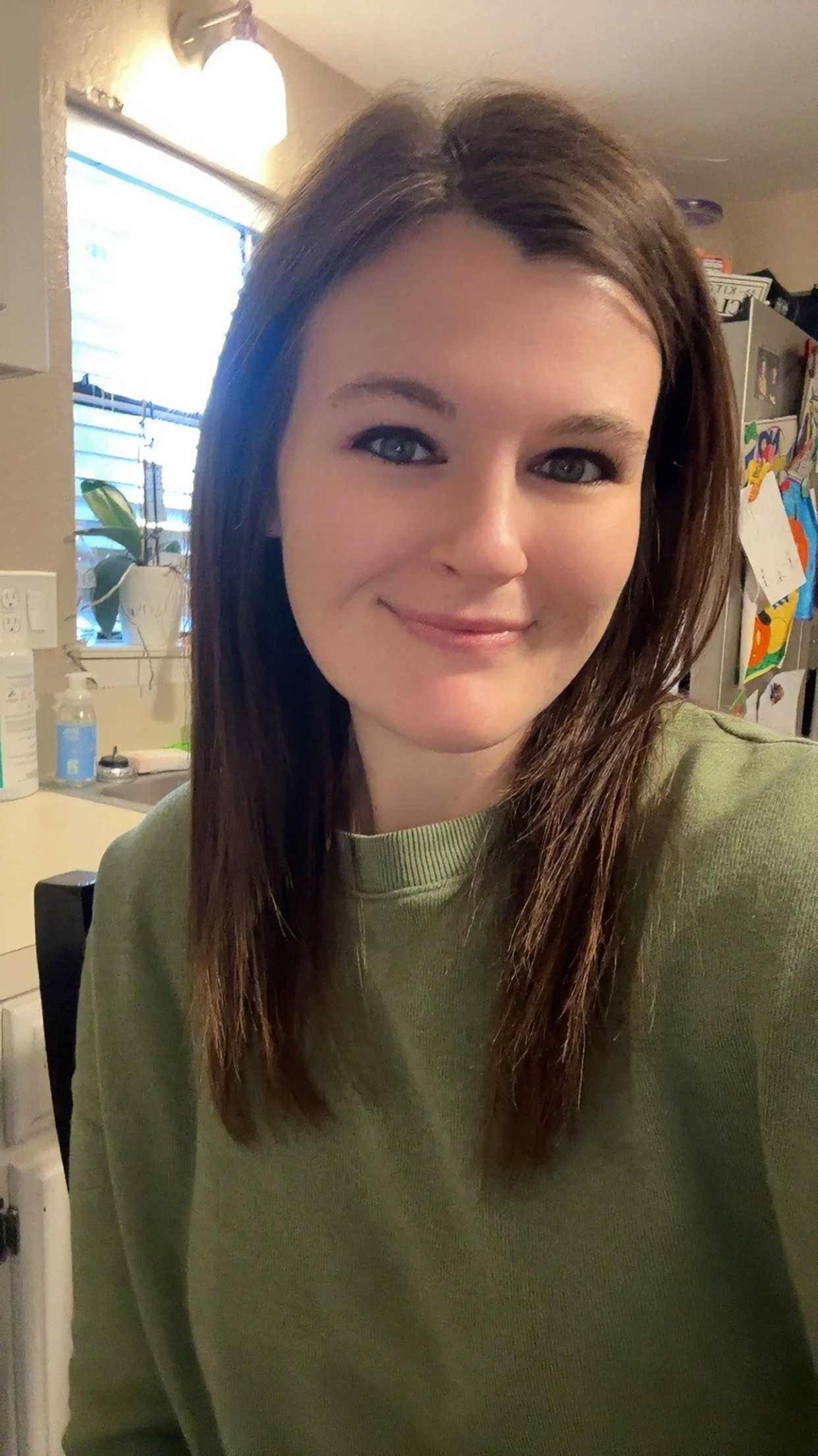 A young woman with shoulder-length brown hair and blue eyes, smiling while taking a selfie in a kitchen.