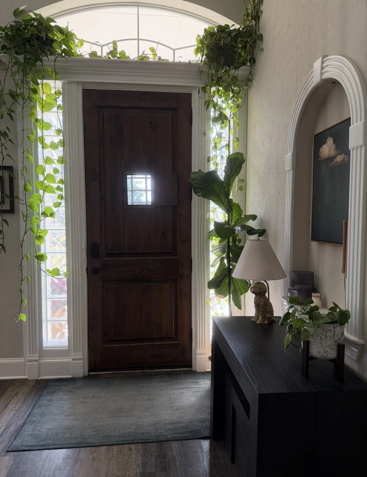 Residential front door with dark wood panels, surrounded by white trim and narrow windows with plants, and decorated interior with a side table, lamp, potted plants, and wall artwork.