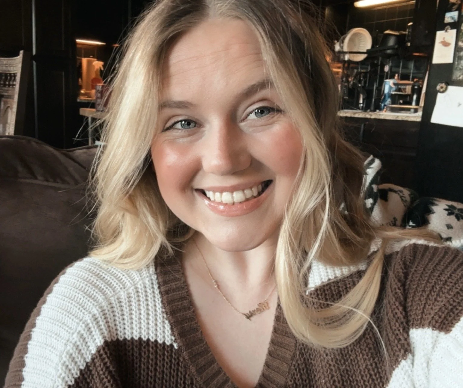 Close-up of a smiling woman with blonde hair, wearing a striped brown and white sweater and a gold necklace, sitting in a cozy indoor setting.