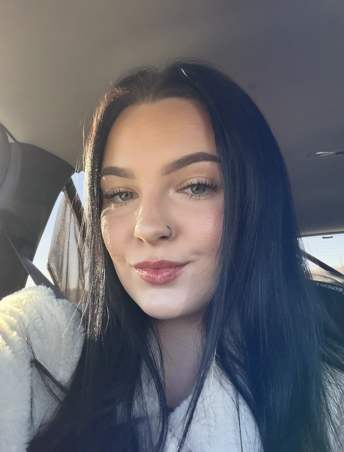 A young woman with black hair, light skin, and blue eyes sitting in a car, taking a selfie. She is wearing makeup and a nose ring, and has a slight smile on her face.
