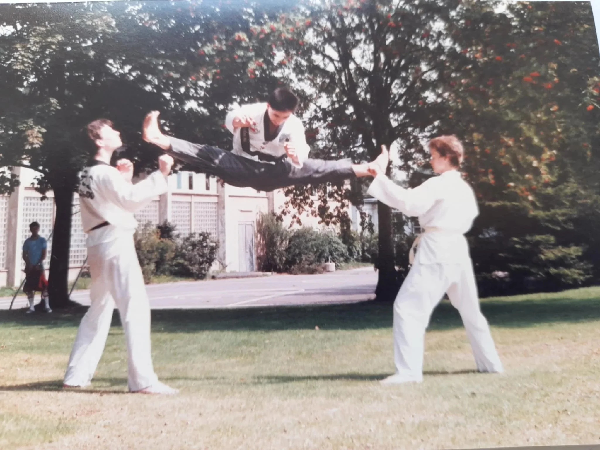 Two martial artists in white uniforms practicing martial arts outdoors, one is airborne performing a kick while the other two support and defend, with trees and a building in the background.