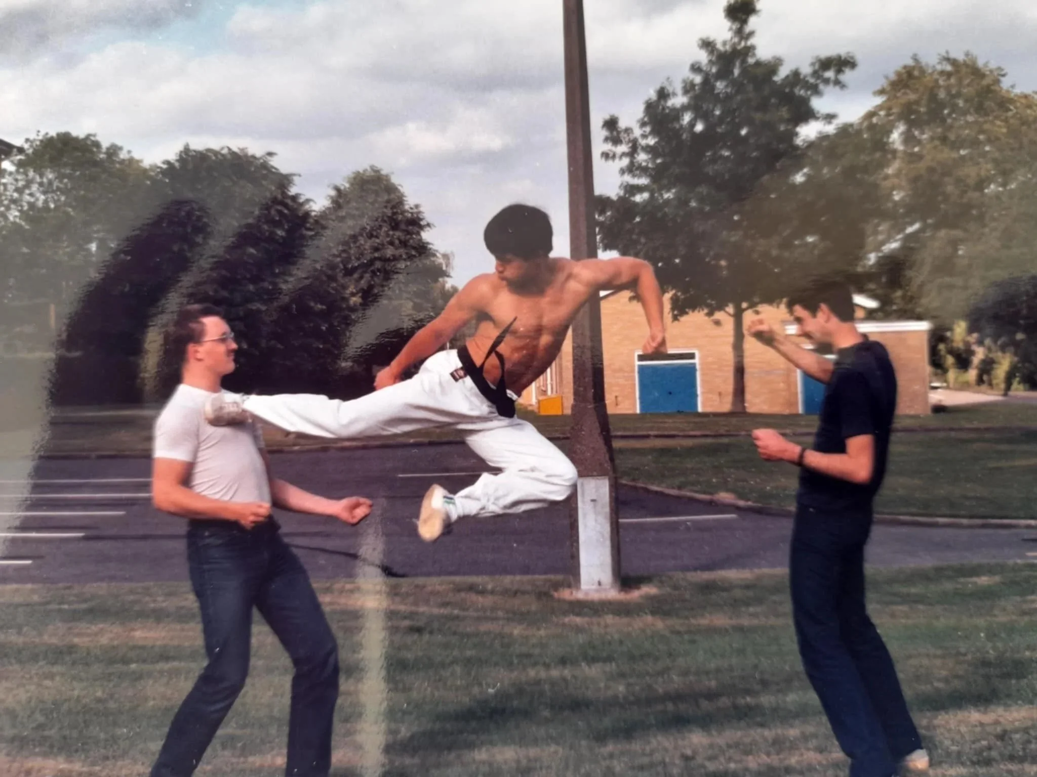 Three young men appear to be having fun outdoors, with one shirtless man mid-air doing a kick, while the other two stand nearby on a grassy area near a parking lot and brick building, with trees and sky in the background.