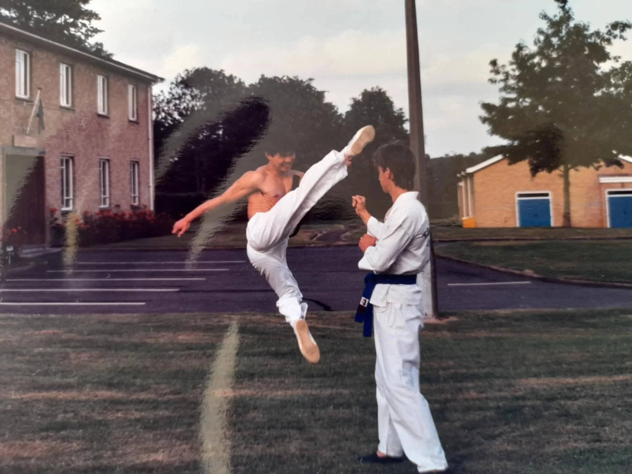 A man performing a high kick during a martial arts demonstration to a person dressed in a martial arts uniform outdoors, with parked cars and buildings in the background.