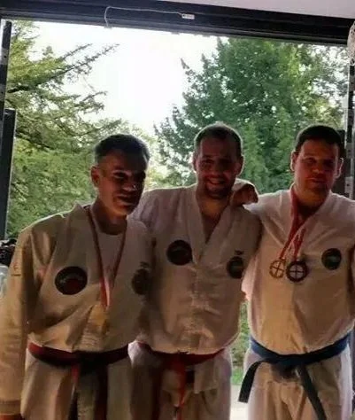 Five young martial artists in gi uniforms and medals standing together inside a training facility, with open doors showing trees outside.