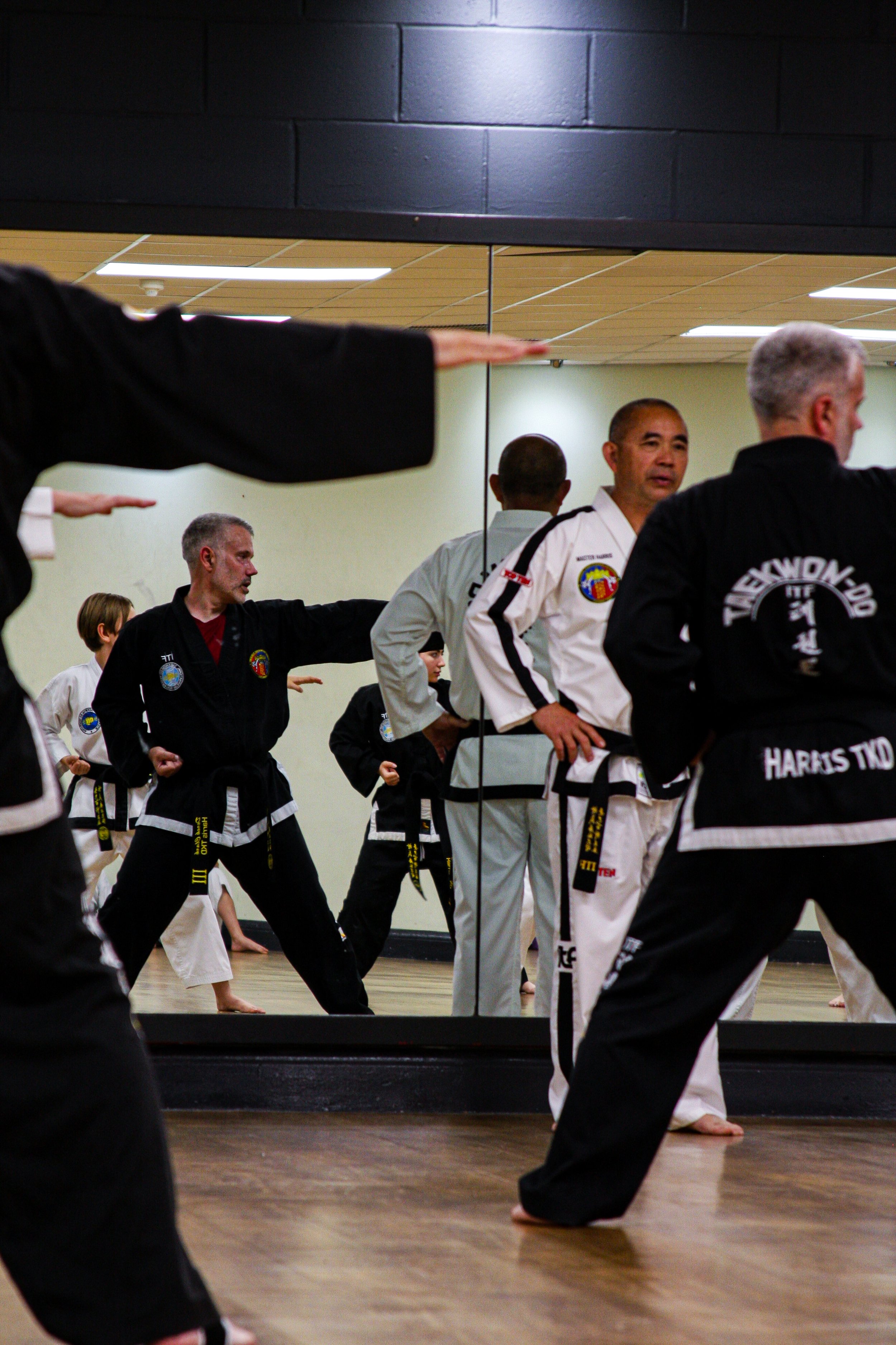 Martial arts students practicing in a studio with a large mirror, wearing martial arts uniforms with patches and belts.