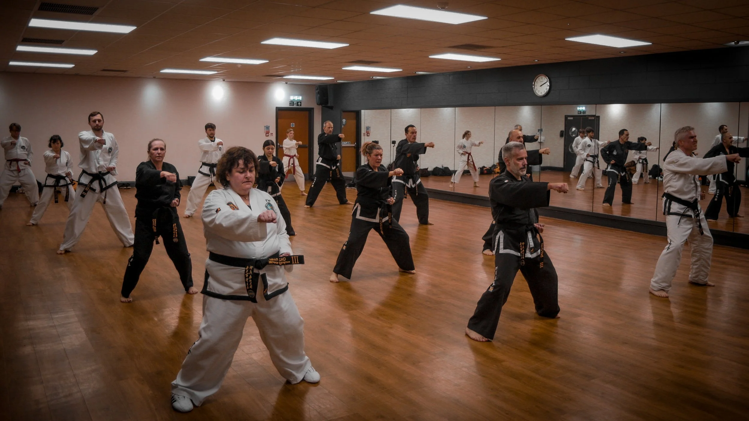 Martial arts class with students practicing punches in a spacious studio with wooden flooring and a large mirror, some students wearing white and others black belts.