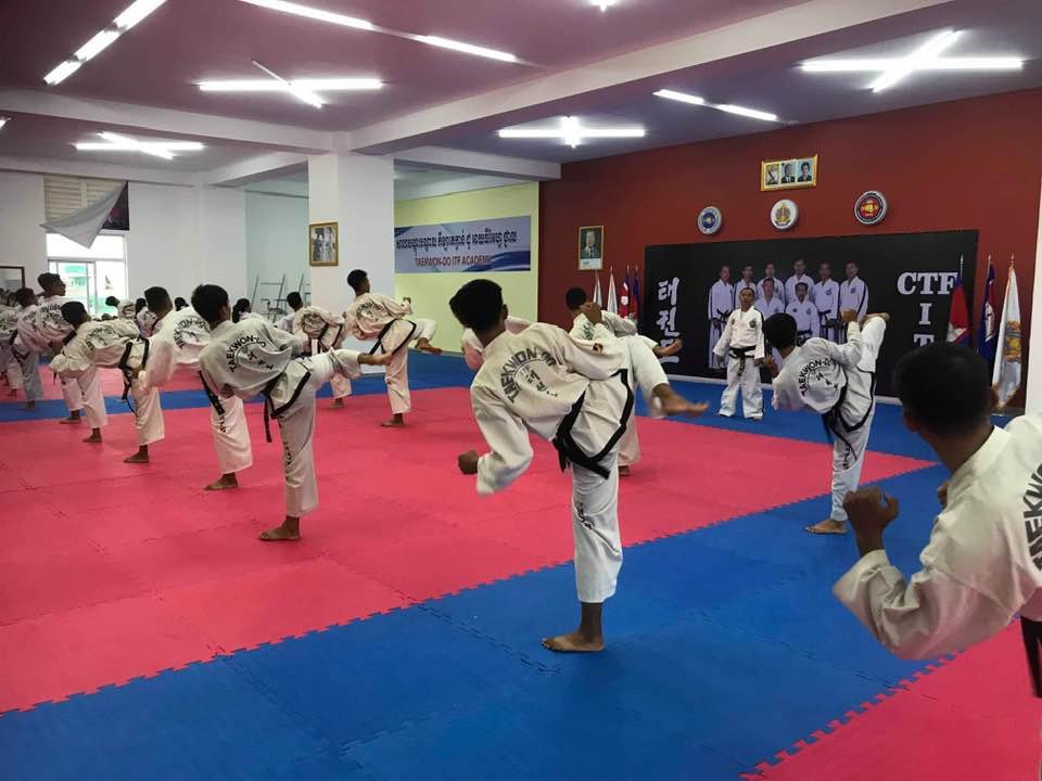 Martial arts students practicing kicking techniques in a dojo, all wearing white uniforms with black belts.
