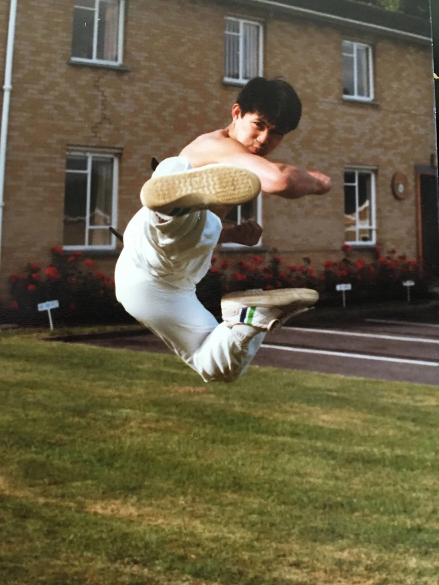 A young man in martial arts attire performing a high kick outdoors in front of a brick building with flowerbeds.