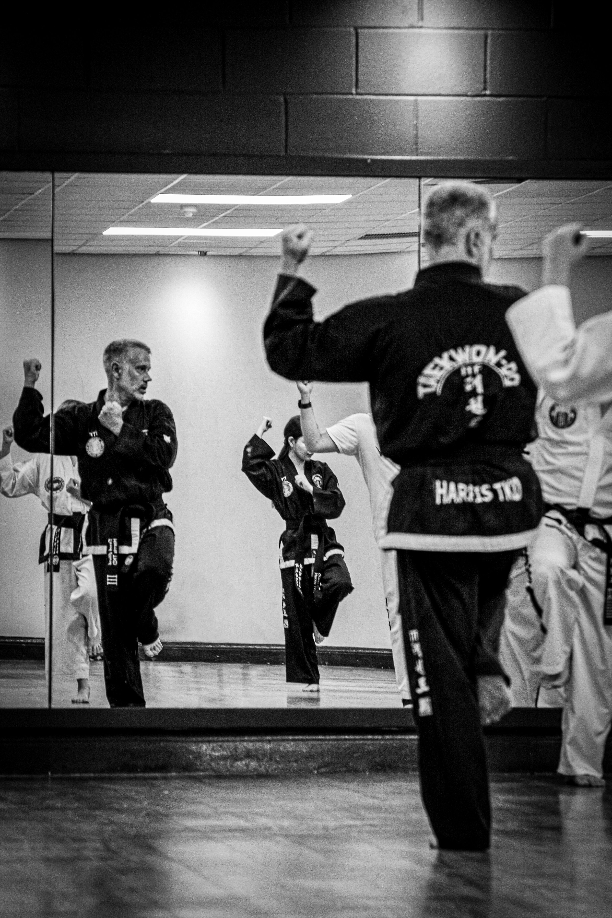 Black and white photo of martial arts students practicing in a studio, with a mirror reflecting their movements, wearing uniforms with patches and belts.