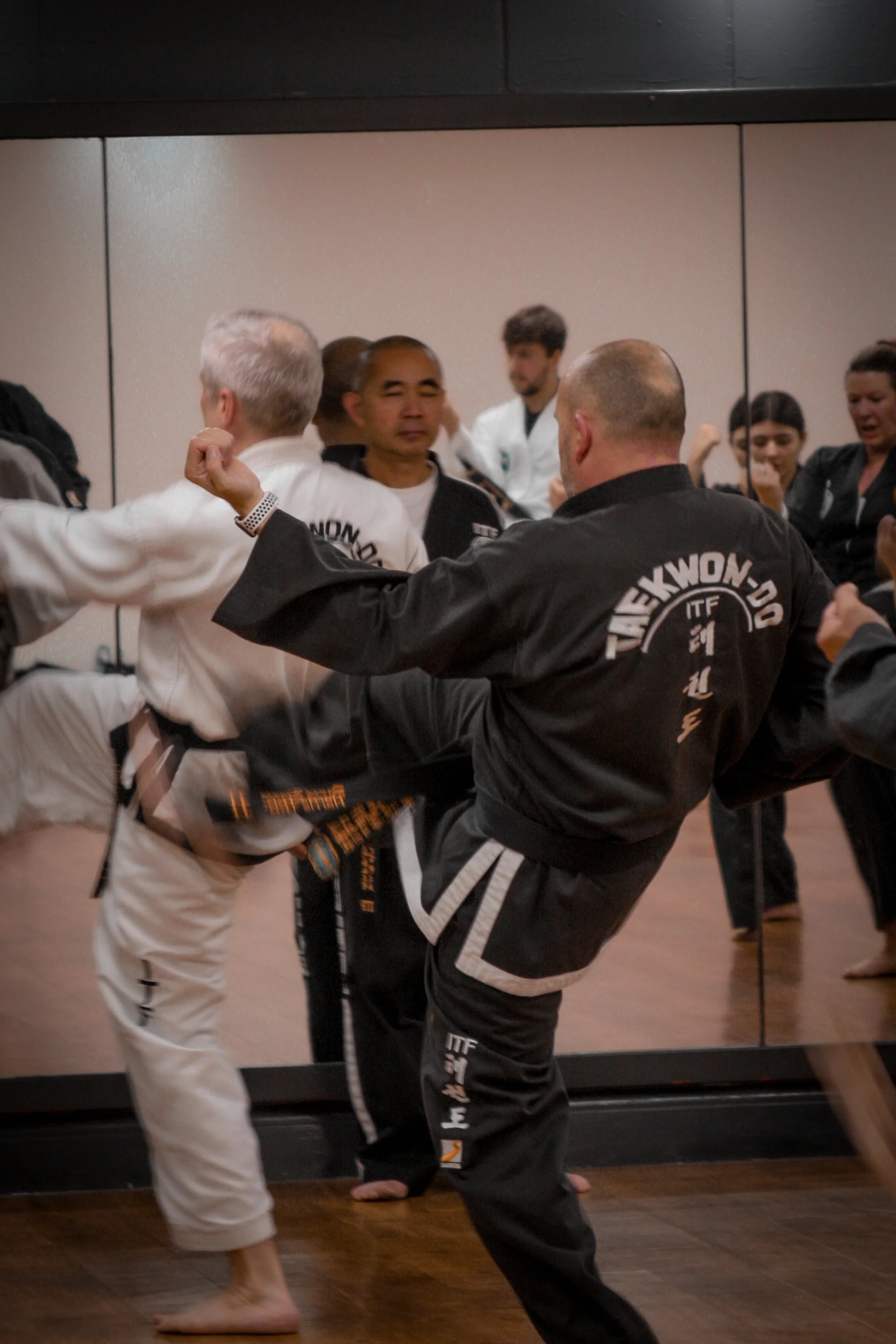 People practicing Taekwondo in a martial arts studio with a mirror. Focus on two individuals executing a high kick, wearing black and white uniforms.