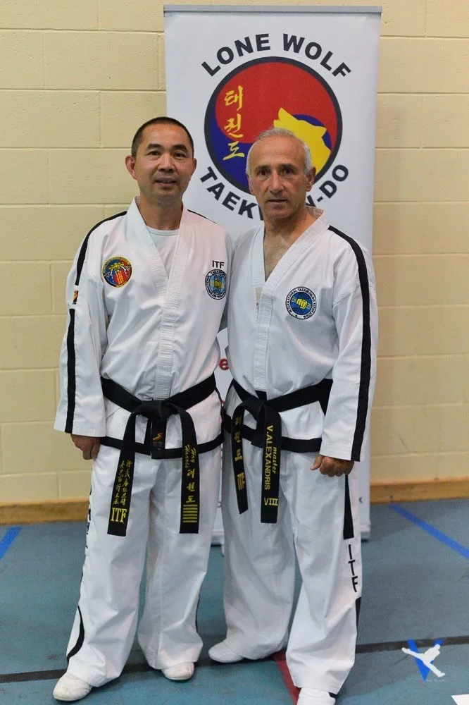 Two men in martial arts uniforms standing in front of a banner that reads "LONE WOLF TAEKWONDO" with a map of South Korea and some Korean text on it.