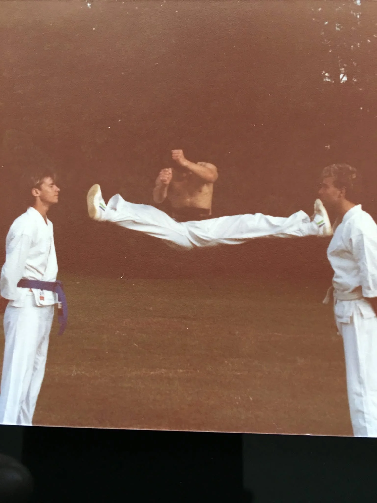 Two martial artists in white gis standing face-to-face, with a shirtless martial artist performing a flying kick between them.