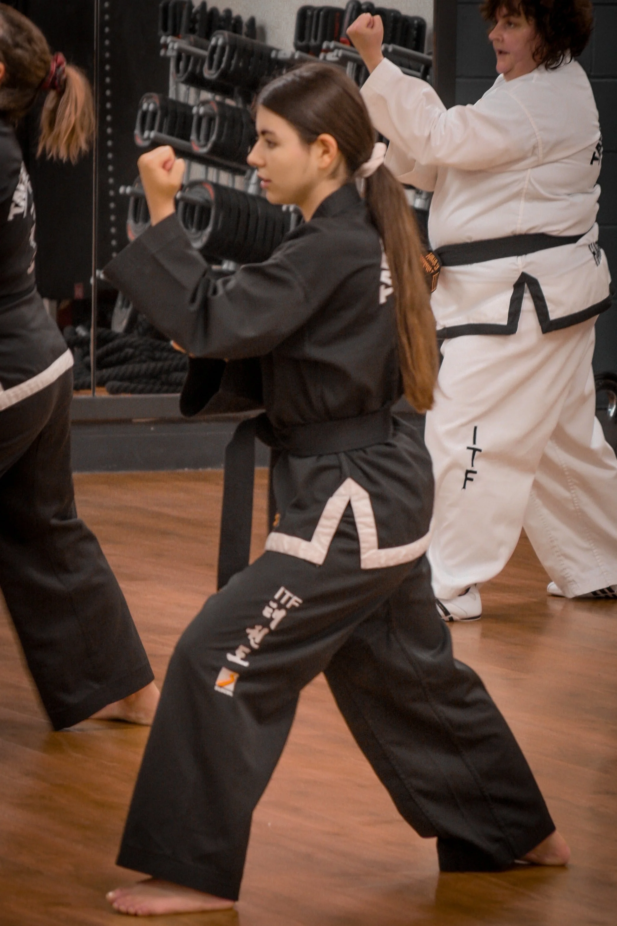 A woman in a black martial arts uniform practicing a fighting stance in a martial arts class, with others in similar uniforms training nearby.
