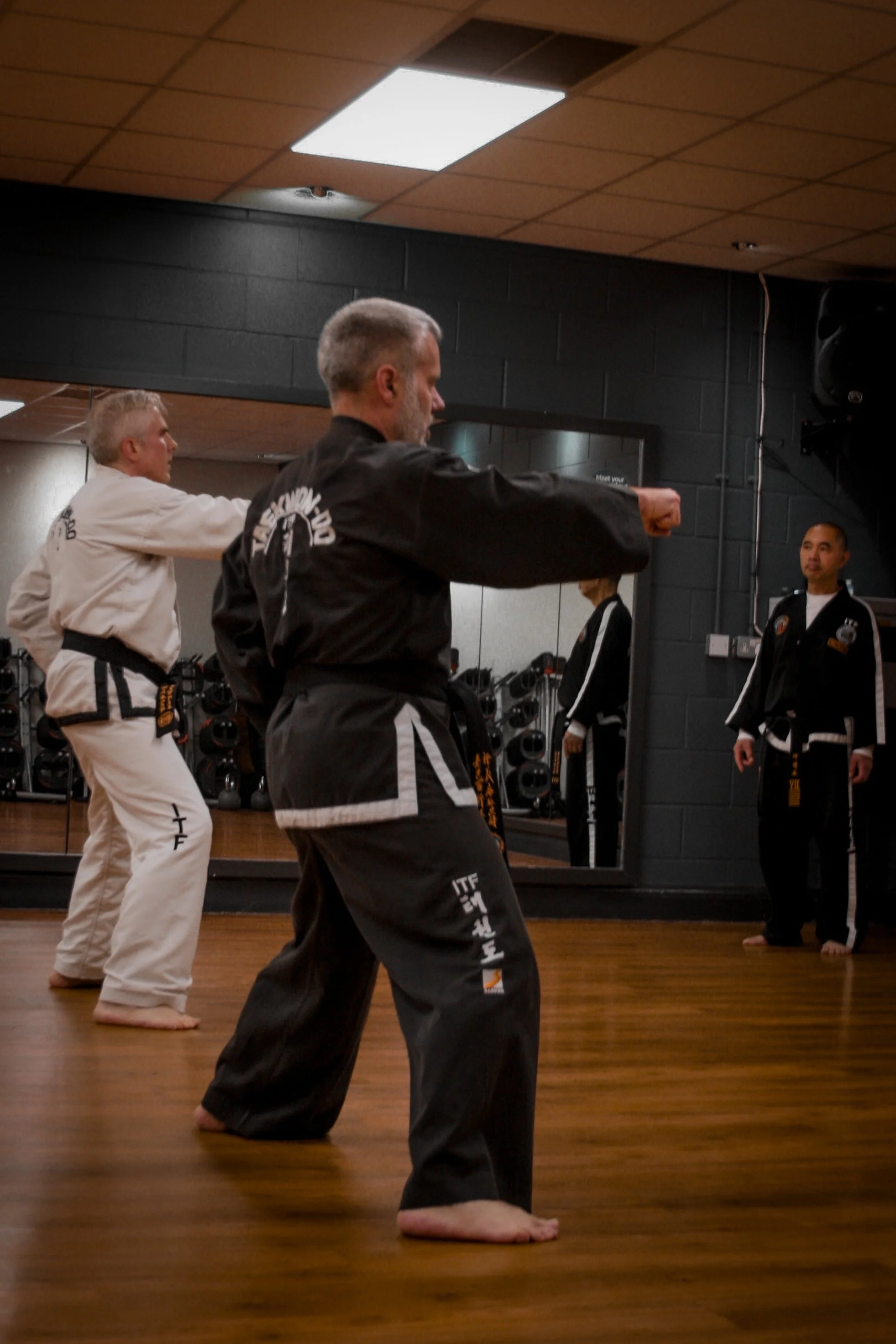 Two martial arts practitioners in black and white uniforms practicing kicks in a dojo, with an instructor observing in front of a mirror.