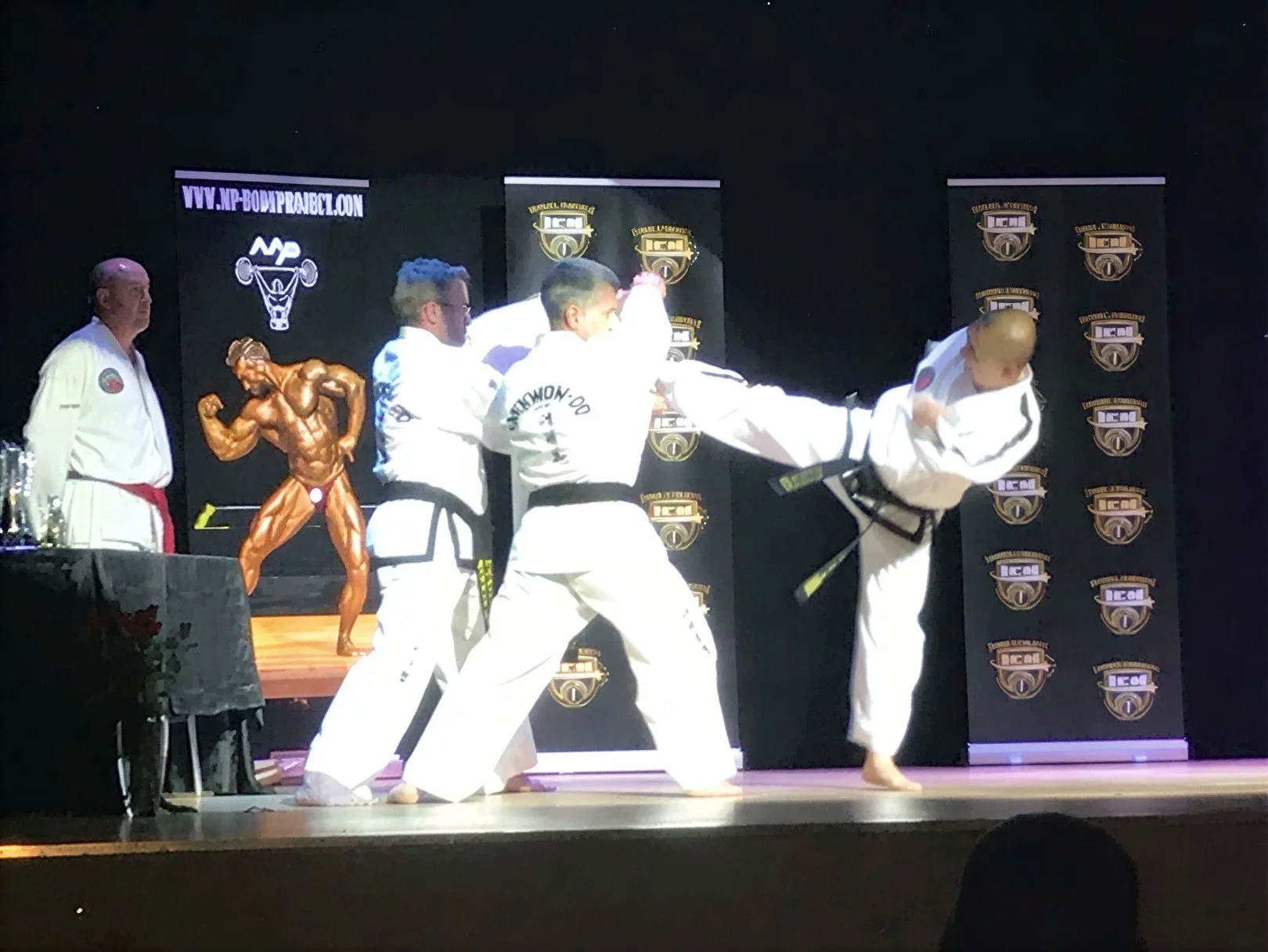 Martial arts demonstration with five practitioners in white uniforms, one executing a high kick, others supporting, on stage with a black backdrop with logos and a table with trophies and a bronze sculpture of a muscular man.