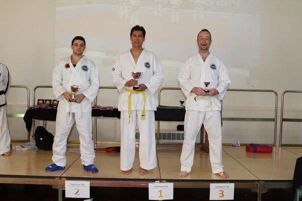Three men in martial arts uniforms standing on a podium, holding trophies, with the middle man in first place, the man on the left in second, and the man on the right in third, in a martial arts competition setting.
