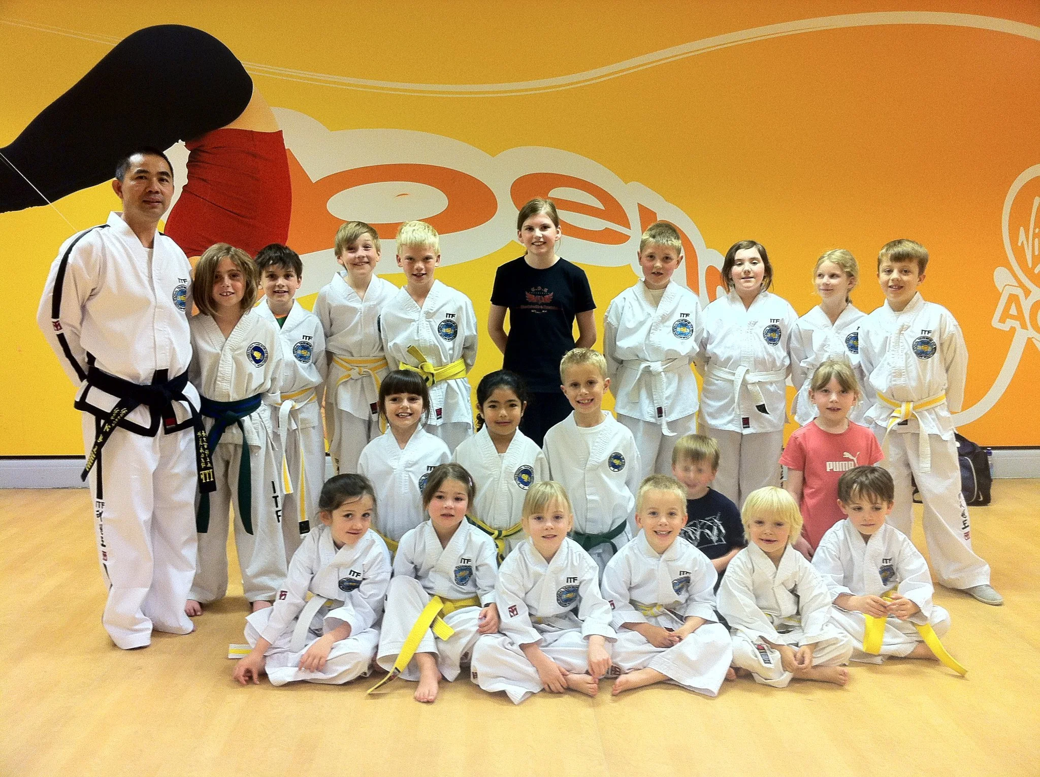 Group of children and their instructor in a martial arts dojo. The children are wearing martial arts uniforms with belts, and the instructor is in a white uniform with a black belt. The background features bright yellow and orange decorations with ma