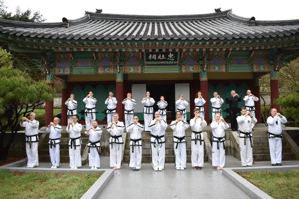 Group of martial artists in uniform practicing martial arts on steps outside a traditional Asian-style building with ornate, colorful roof and wooden beams.