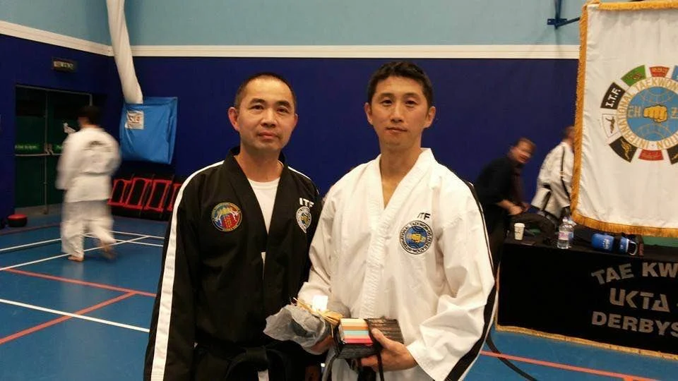 Two men in taekwondo uniforms posing indoors, one in a white uniform and the other in a black jacket, with martial arts awards or certificates.