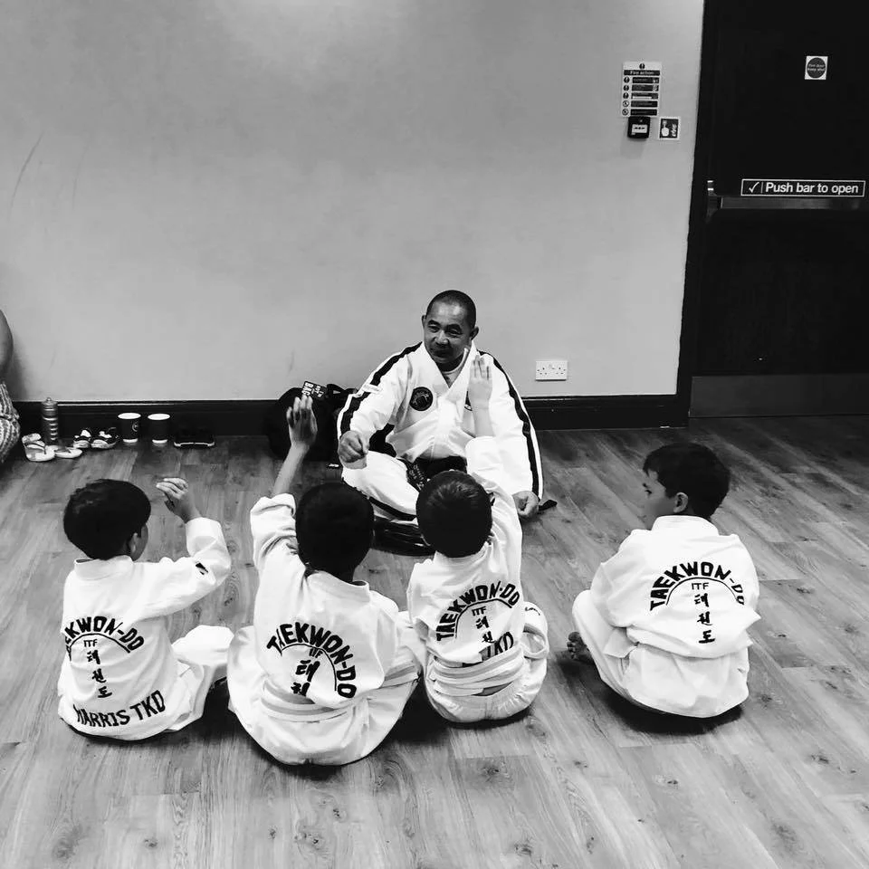 A Taekwondo instructor demonstrating techniques to four children seated on the wooden floor, all wearing Taekwondo uniforms with the logo on the back, in a martial arts studio.