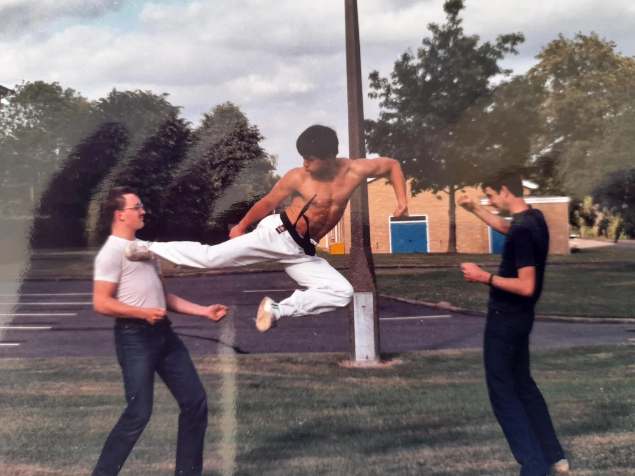 Two young men are practicing martial arts outdoors on a grassy area, with a third man mid-air delivering a kick between them. The man on the left wears glasses and a white t-shirt, the man in the middle is shirtless, and the man on the right is in a 