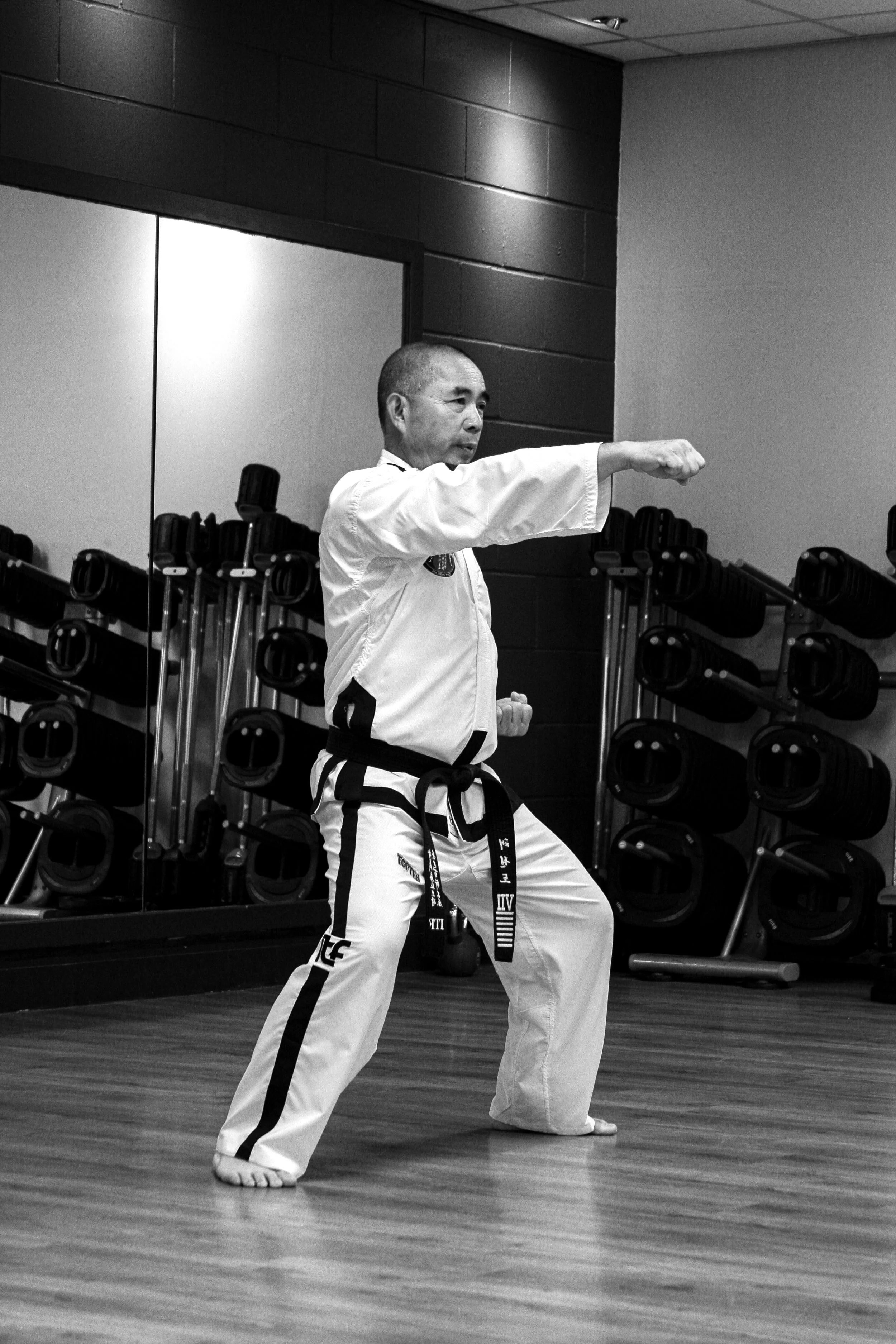 A martial artist practicing a punch in a gym, wearing a traditional uniform with a black belt, in front of mirrors and workout equipment.