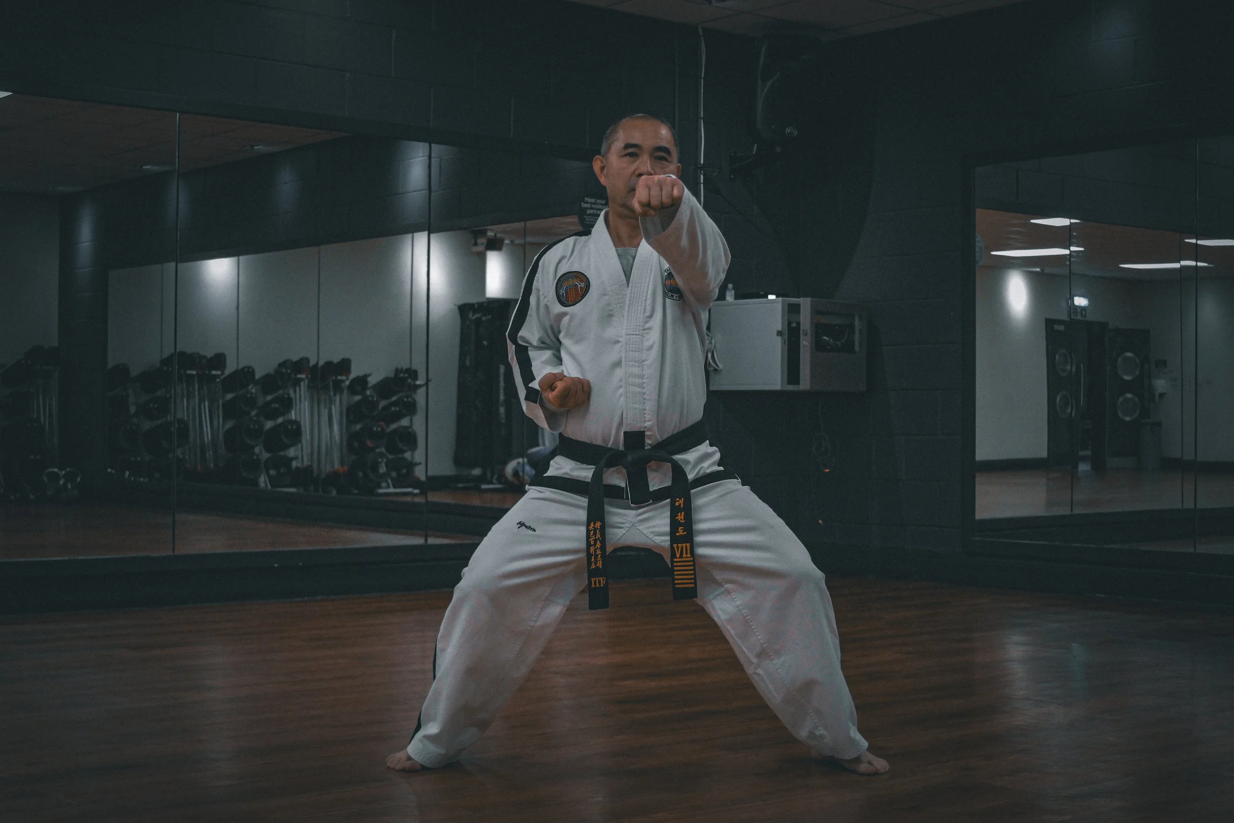 A man in a martial arts uniform performing a stance or punch in a dojo or gym with mirrors and equipment in the background.