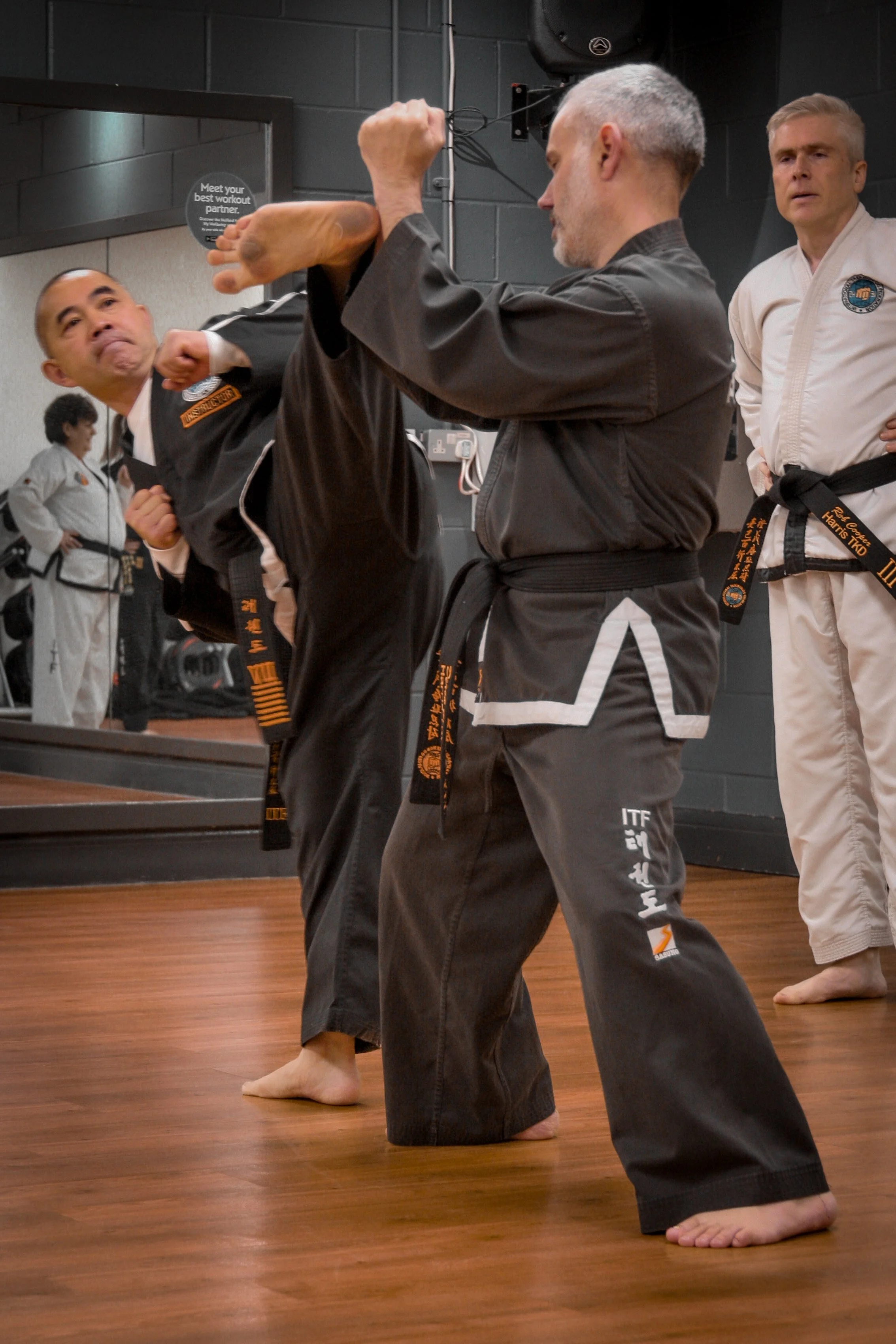 Martial arts class in progress with two men practicing kicking techniques while an instructor observes, all dressed in martial arts uniforms.