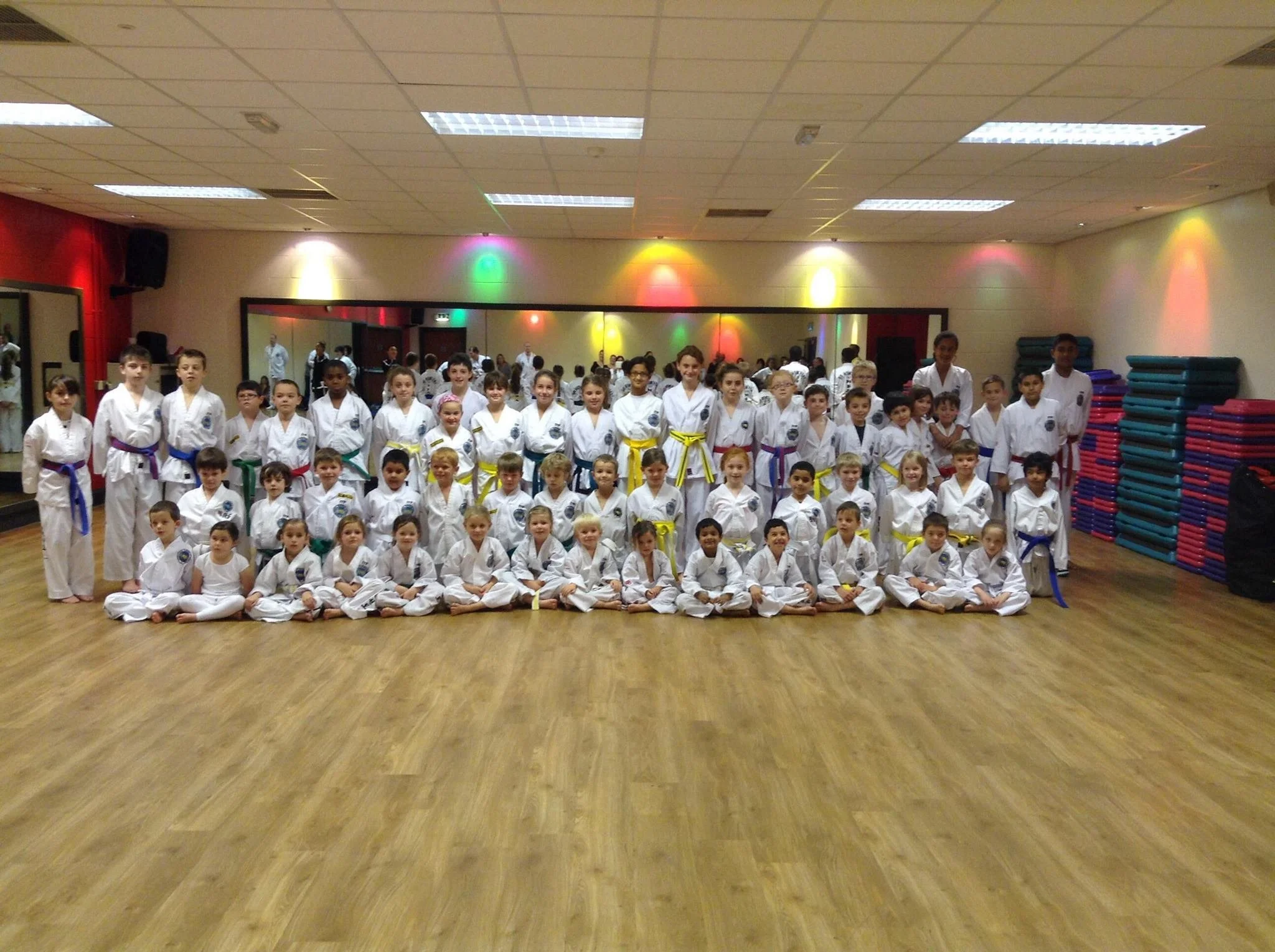 Group of children and instructors in a martial arts dojo, wearing martial arts uniforms with colored belts, posing for a group photo.