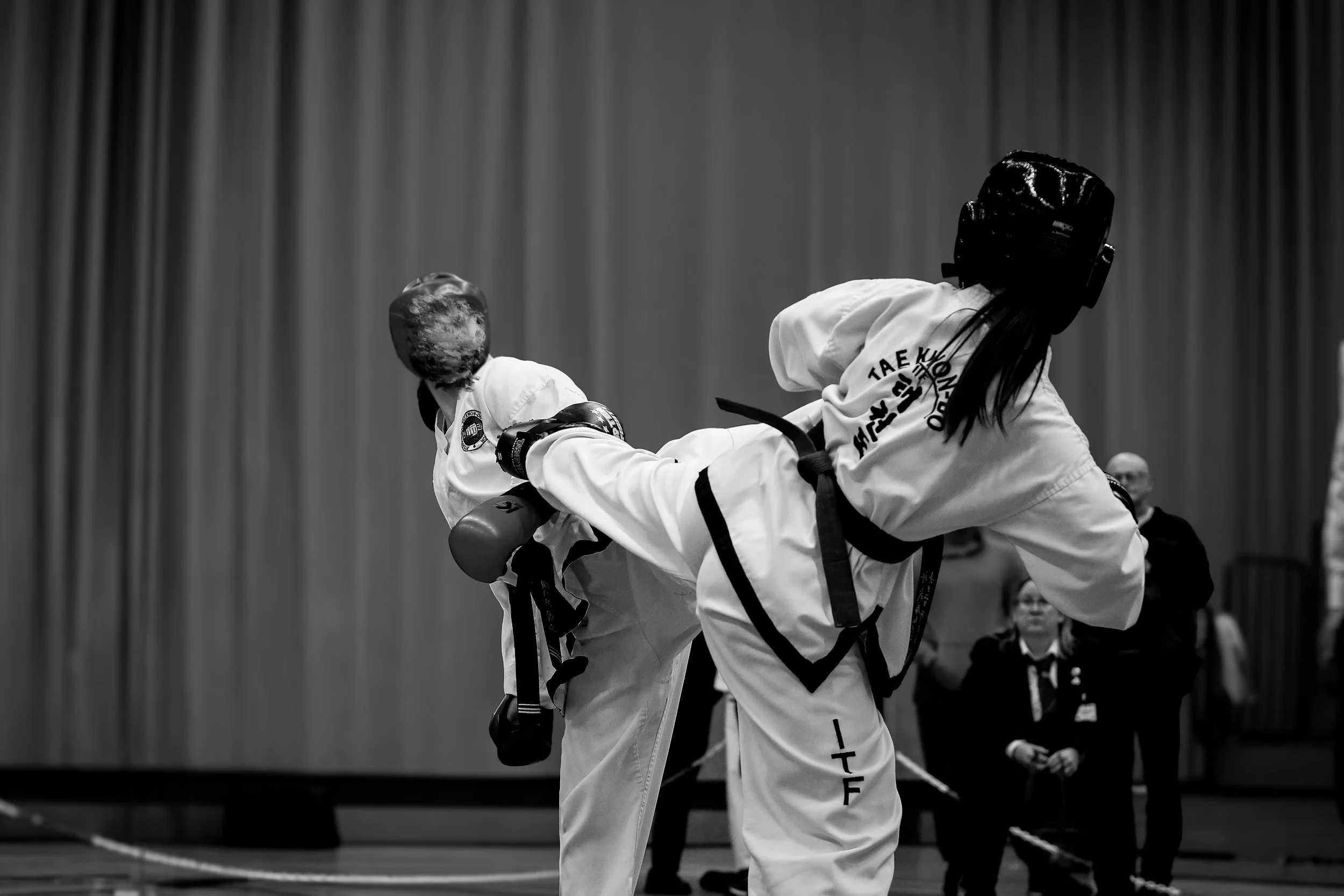 Two taekwondo practitioners in black belts practicing in a gym, wearing protective gear.