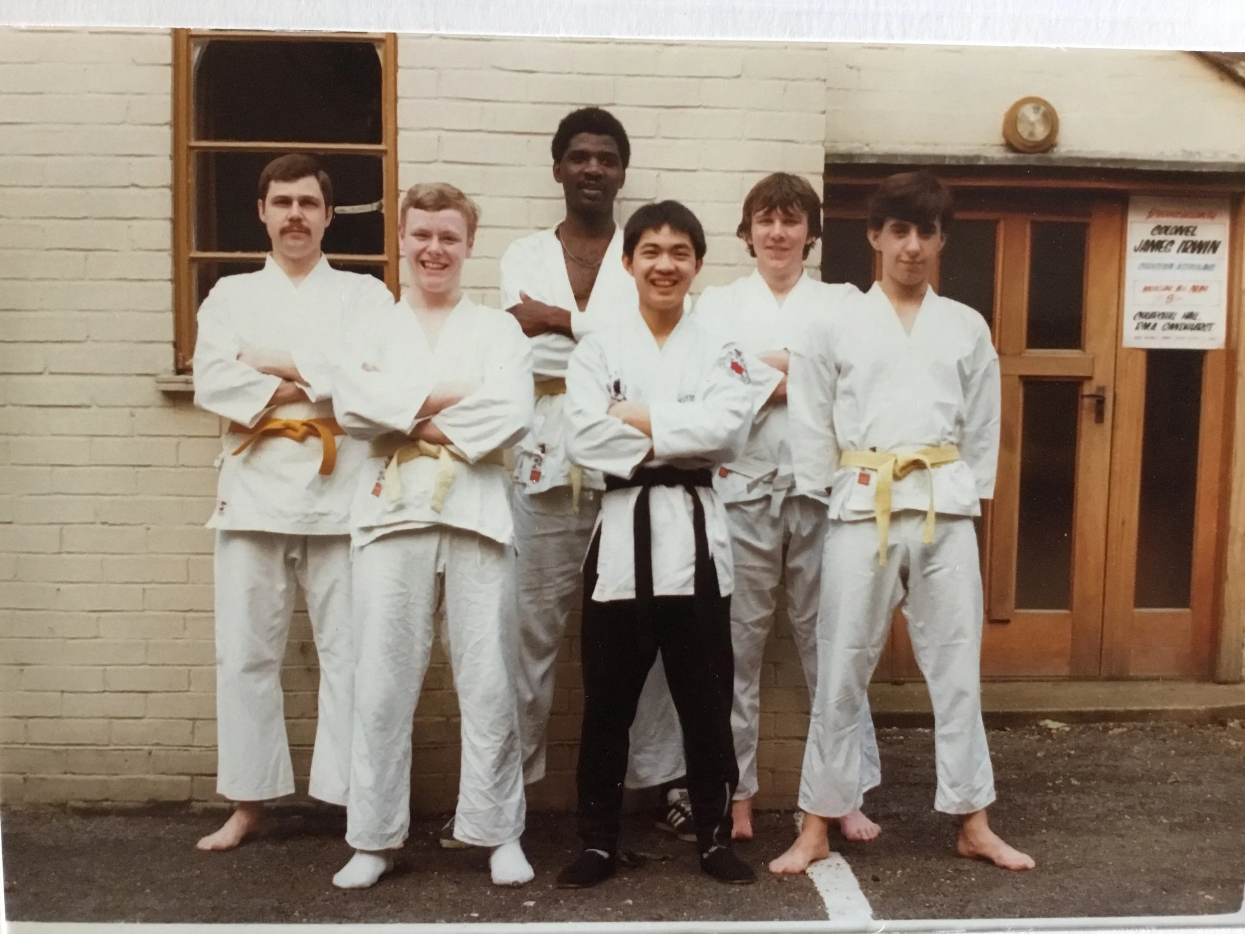 A group of six young men and boys in martial arts uniforms with belts, standing in front of a brick building. They are posing with crossed arms, some smiling, with a doorway and a window behind them.