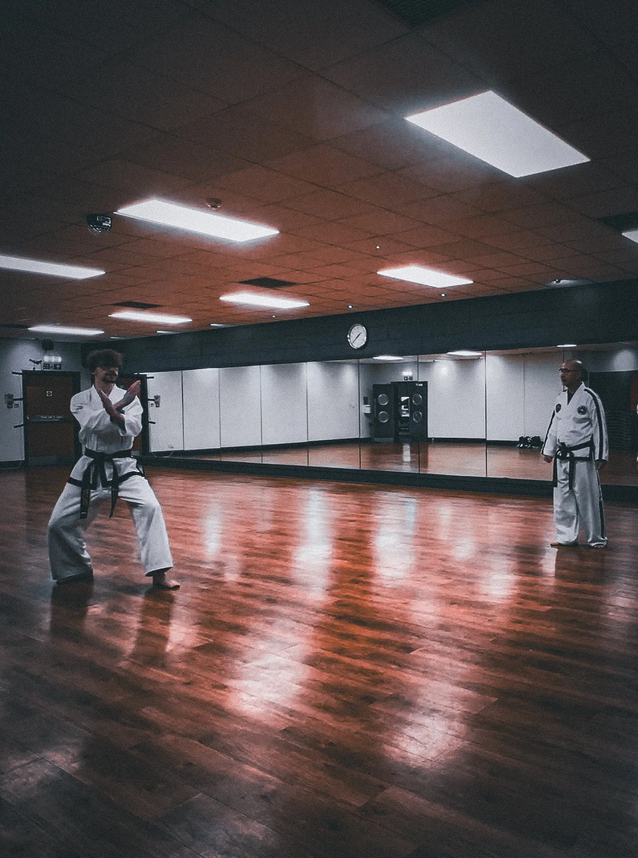 Two individuals practicing martial arts or taekwondo in a spacious, empty training room with wooden floors and a large mirror on the wall.