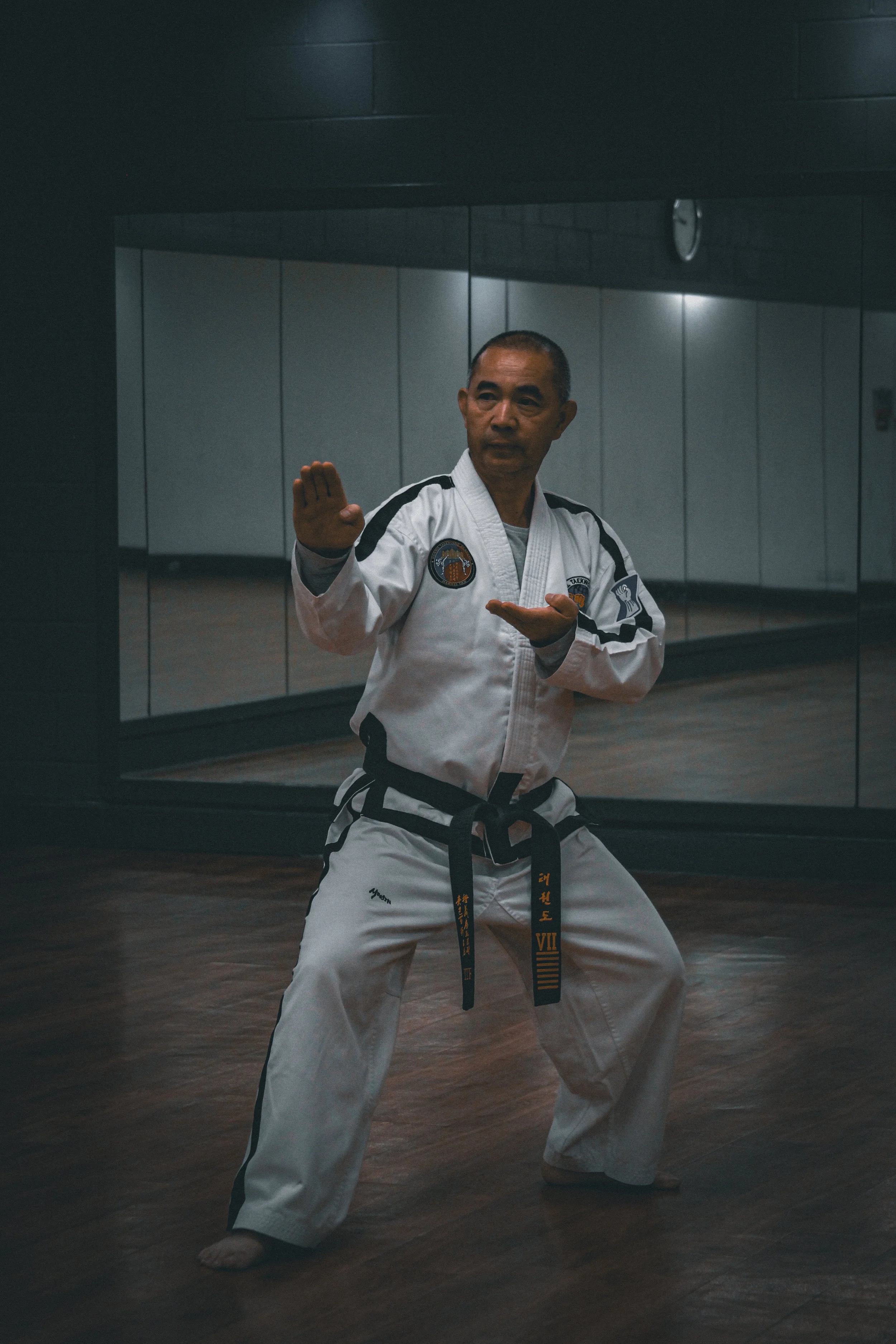 A man practicing martial arts in a gym, dressed in a white uniform with a black belt, standing in a ready stance in front of a mirror.